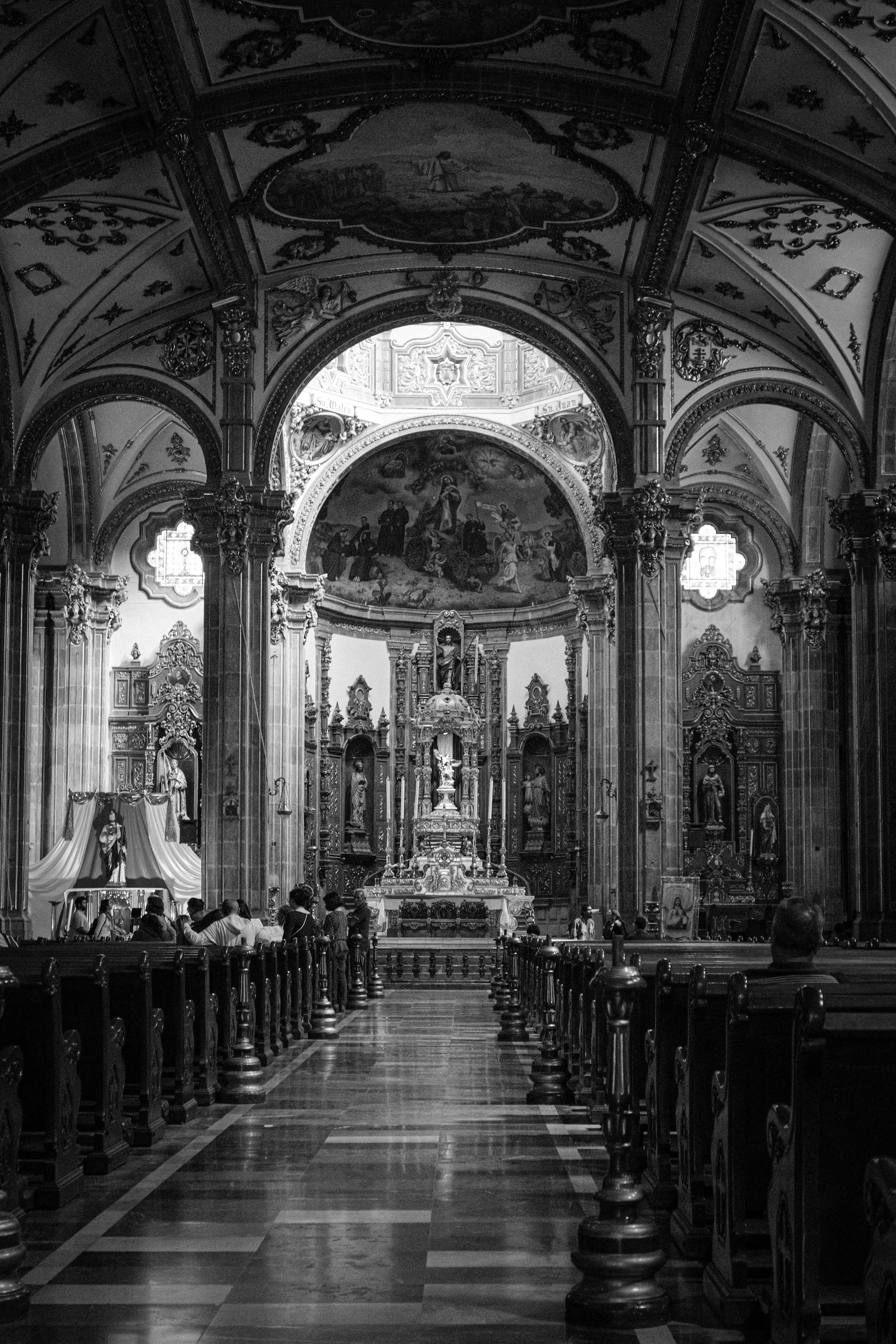 Interior of an ornate church with high arched ceilings, detailed artwork, and a central altar. Several people are seated in pews, and the church features elaborate woodwork and religious icons.
