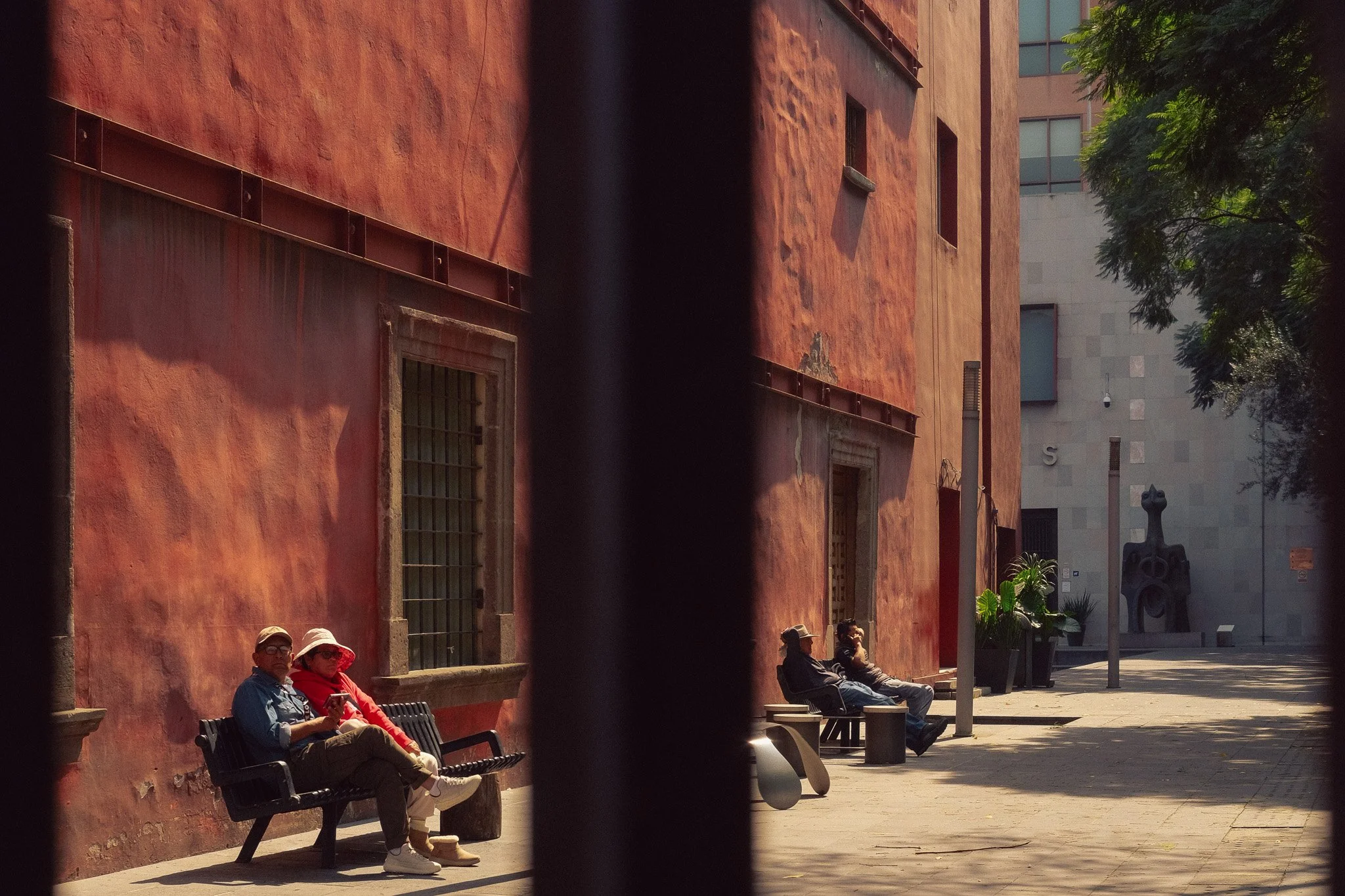 People sitting on benches along a sidewalk next to a red brick building, with a modern sculpture and trees in the background.