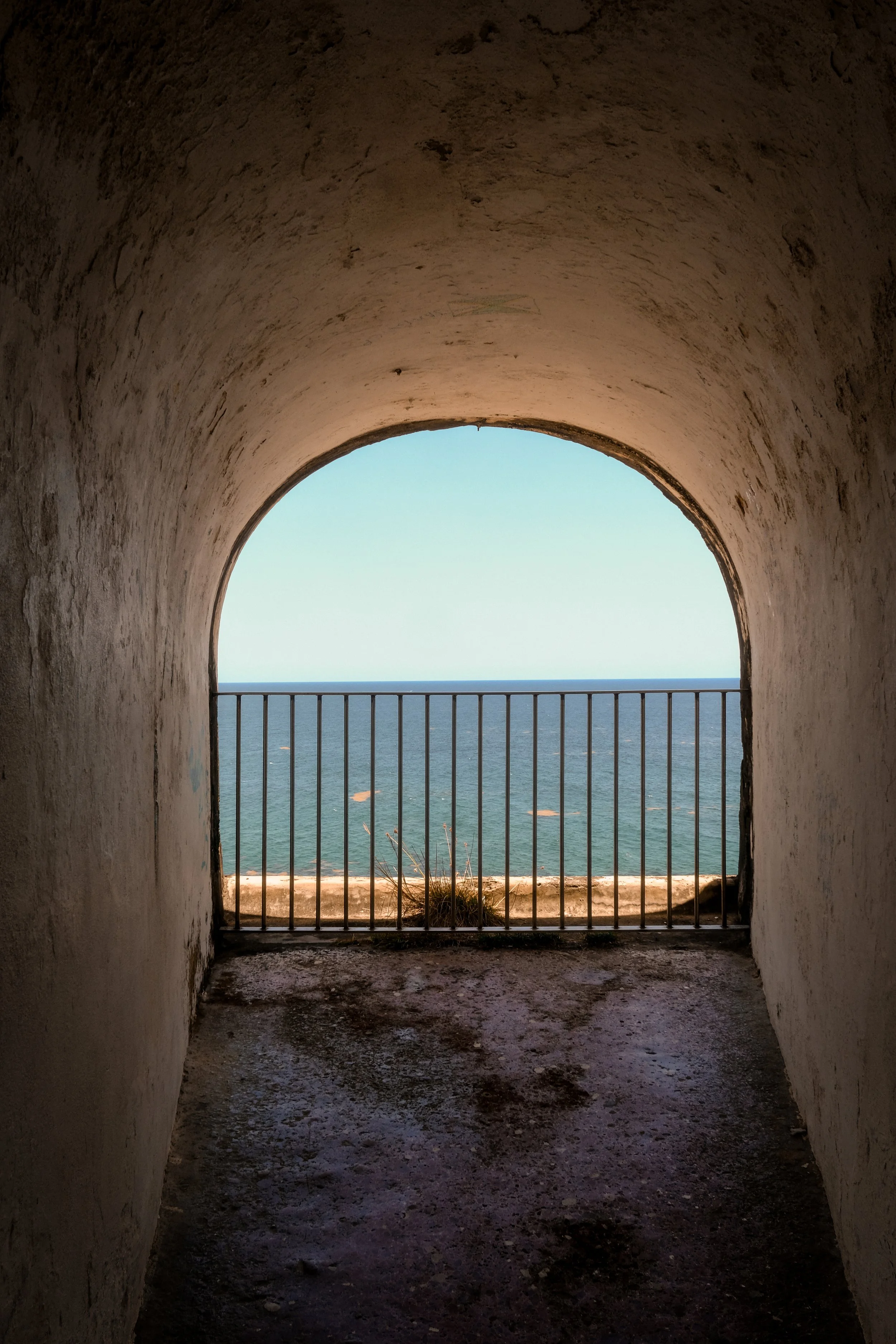 View of ocean through a curved, tunnel-like opening with a metal railing, weathered white walls, and a dark, rough floor.