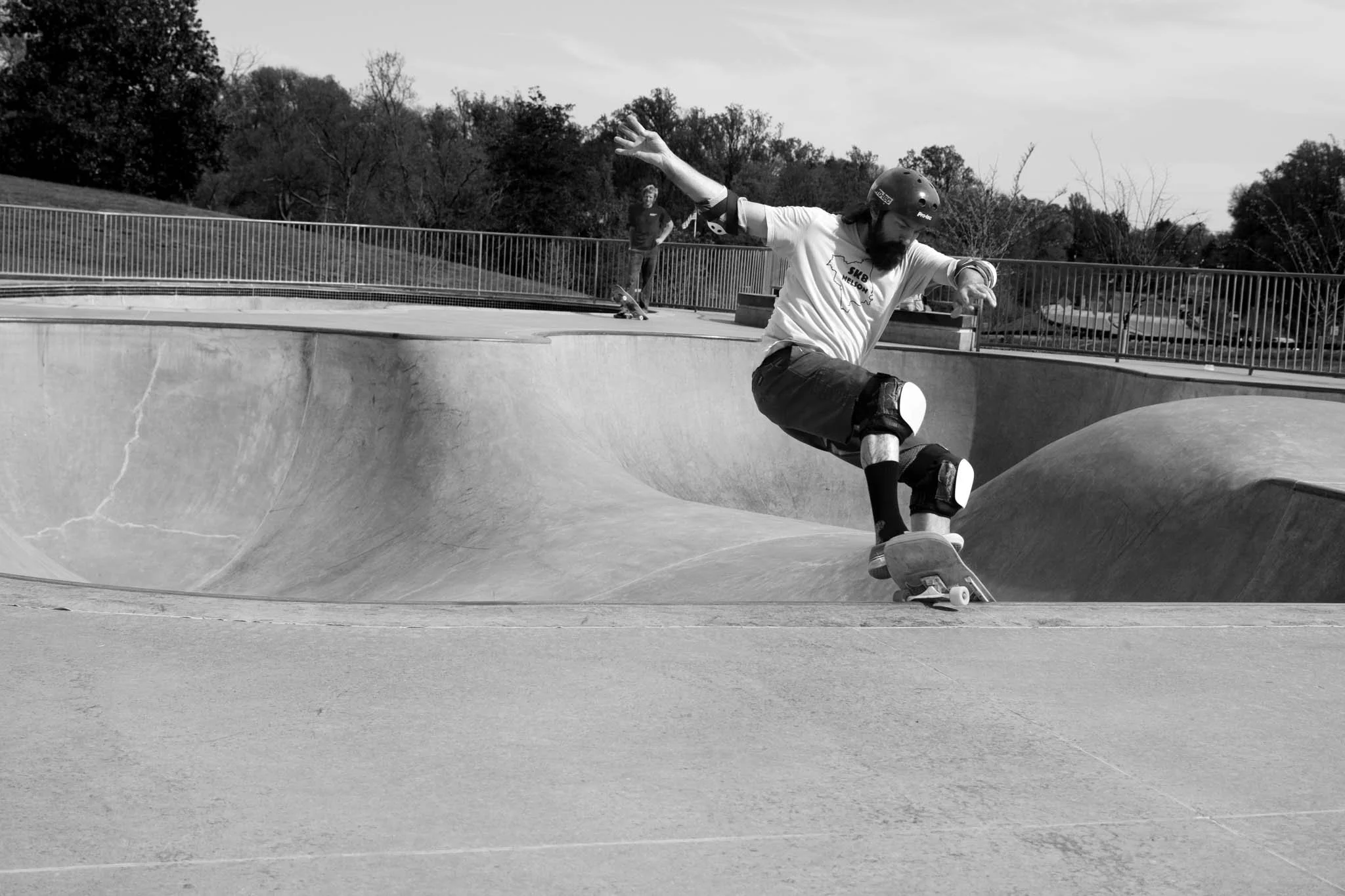 A man skateboarding at a skate park, wearing a helmet, protective pads, and casual clothing, with trees and another person standing in the background.