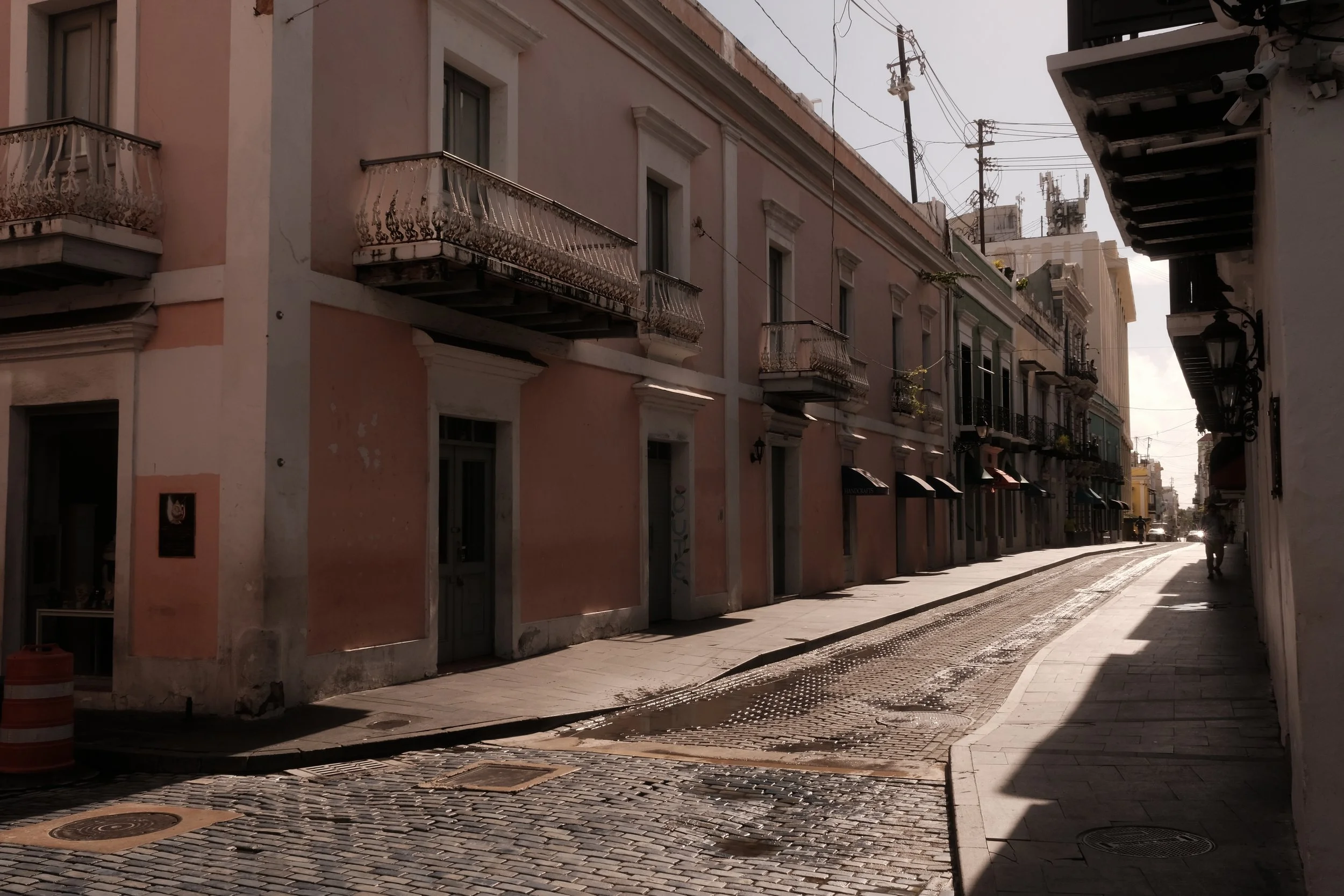 Empty cobblestone street in a historic neighborhood with pastel-colored buildings, some with balconies, and a person walking in the distance.