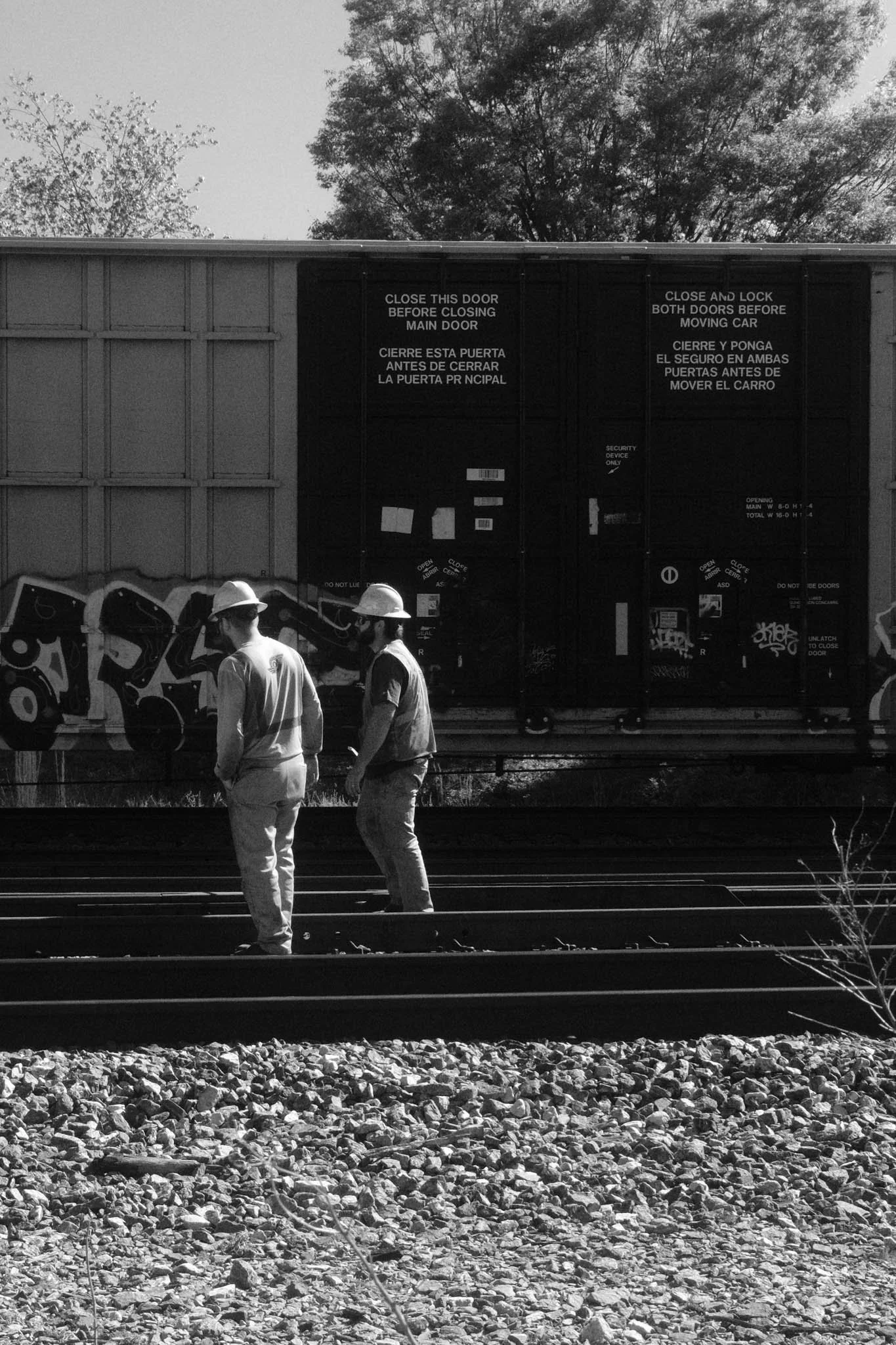 Two construction workers wearing safety helmets and vests standing on railway tracks in front of a train car with graffiti, trees in the background.