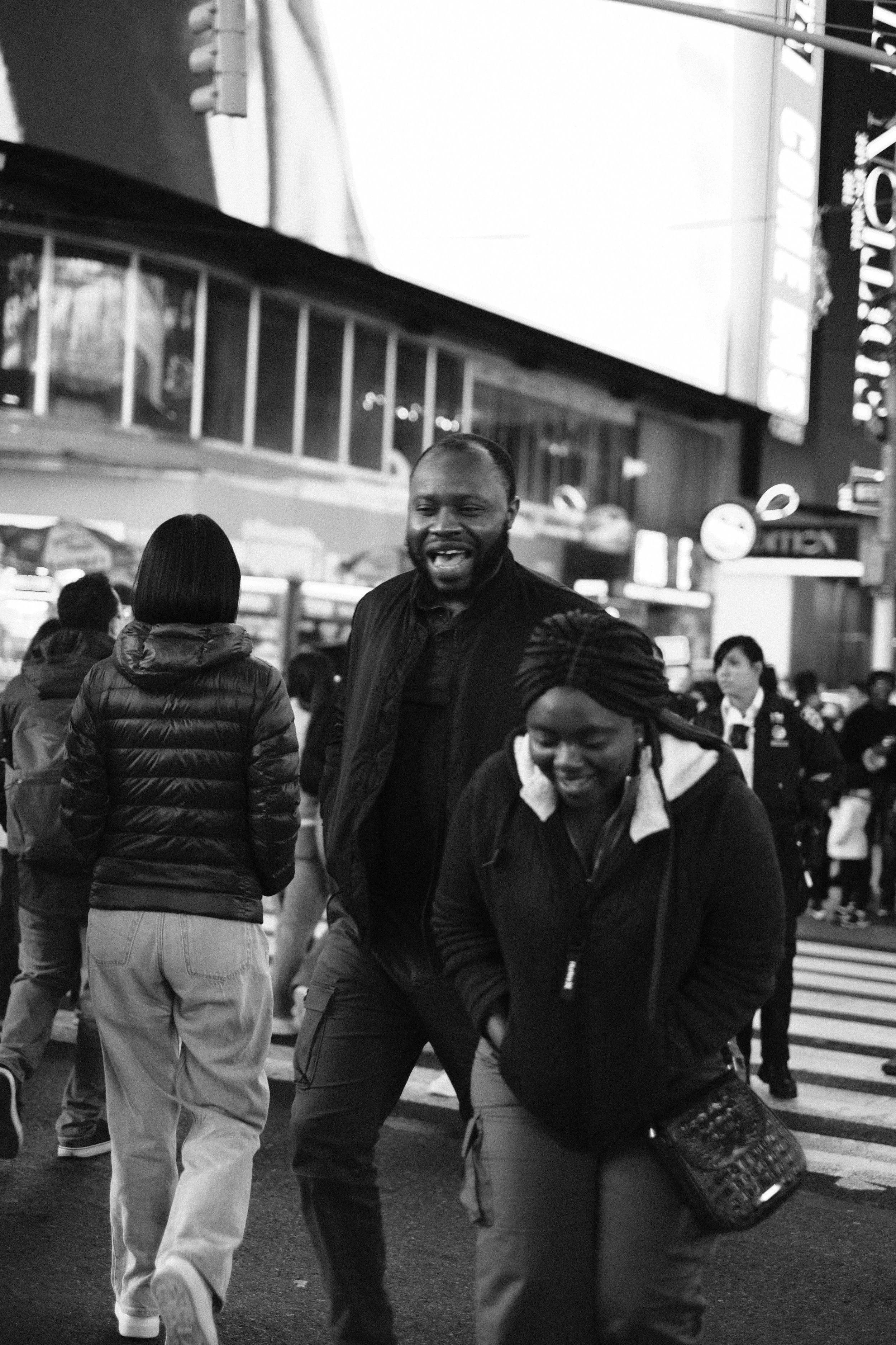 A group of people crossing the street in a city at night, with a large illuminated billboard and signs in the background, in black and white.