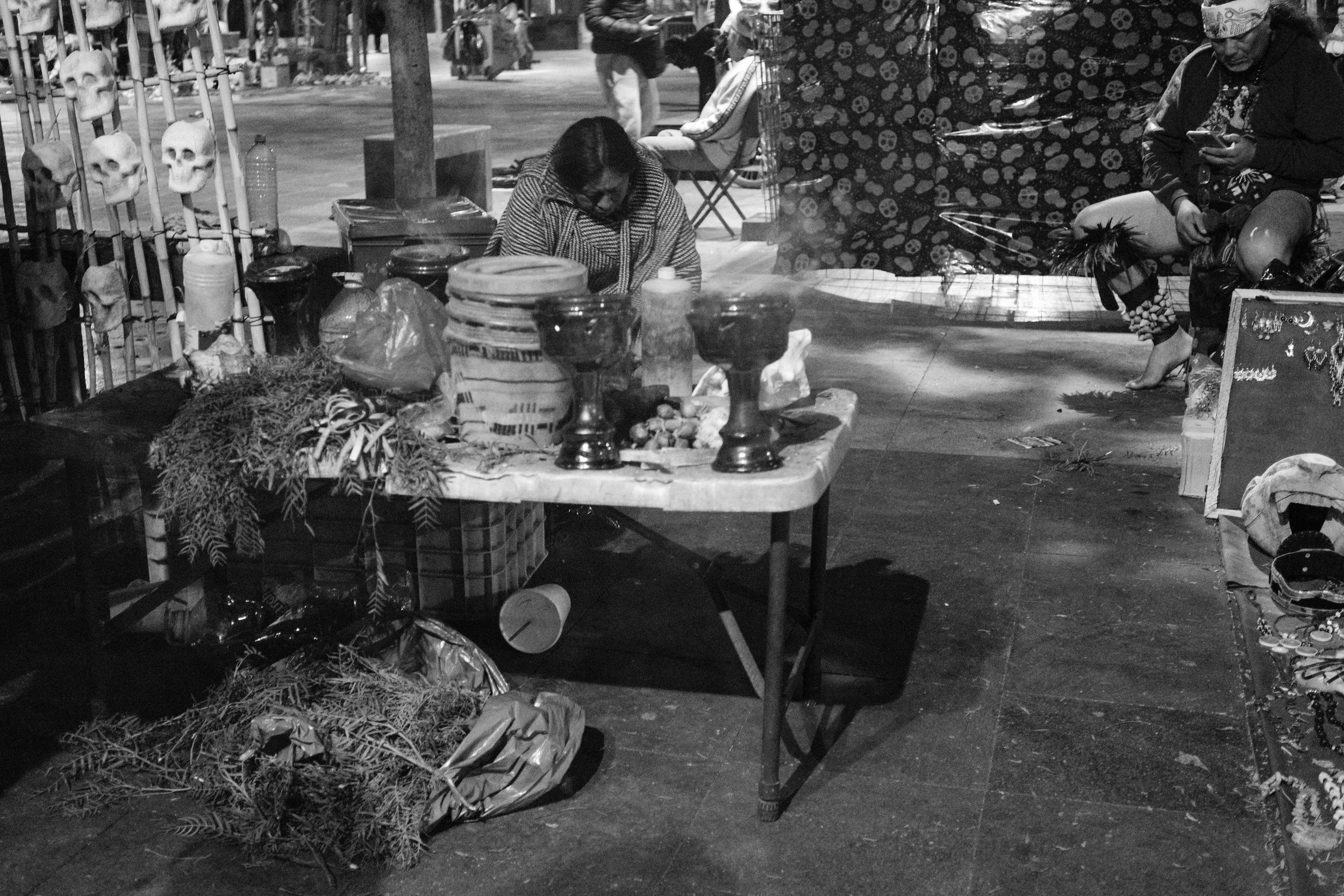 Street market stall at night with various items, a table with vases, jars, and food, surrounded by people and skull decorations.