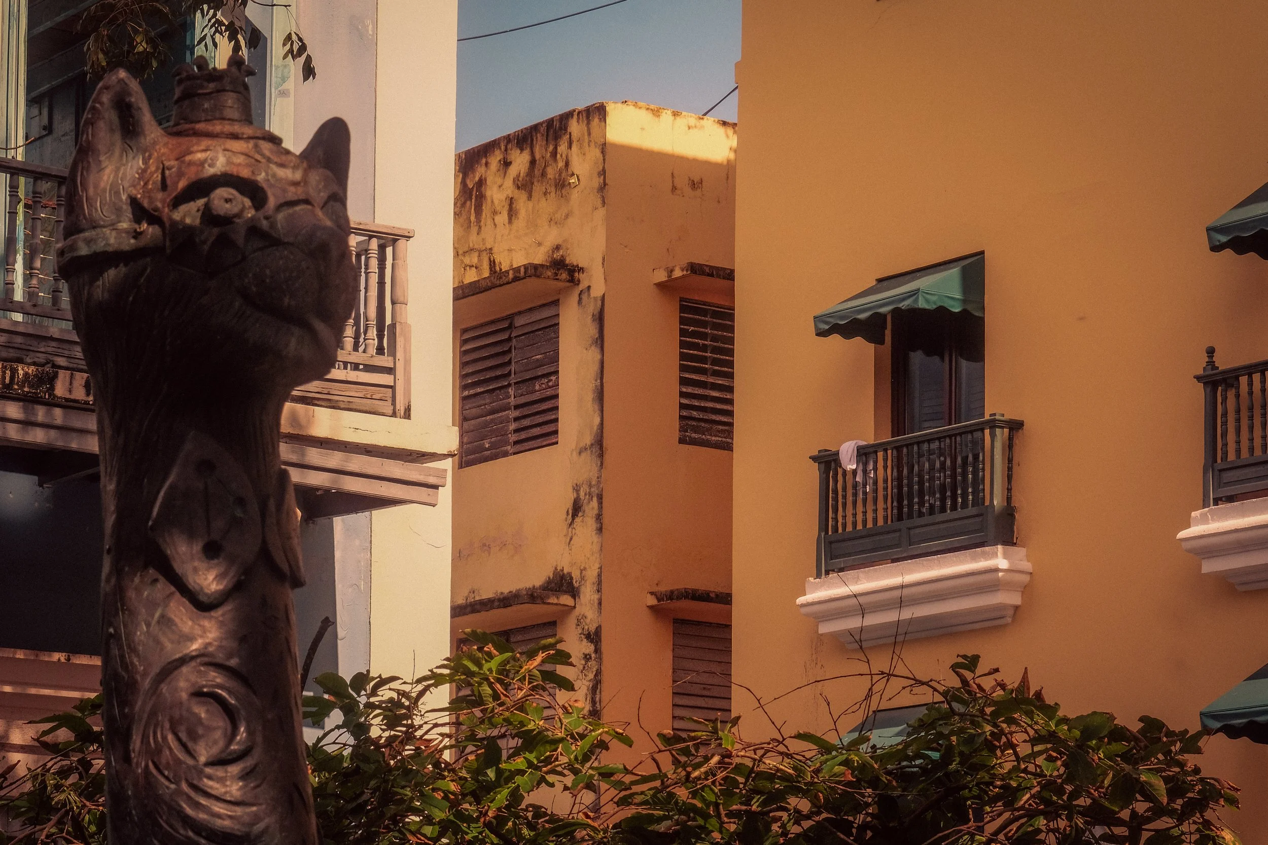 A tall carved wooden figure resembling a cat or feline standing in front of a colorful building with multiple balconies. Some window shutters are closed, and one balcony has a towel hanging. The building shows signs of weathering and aging.