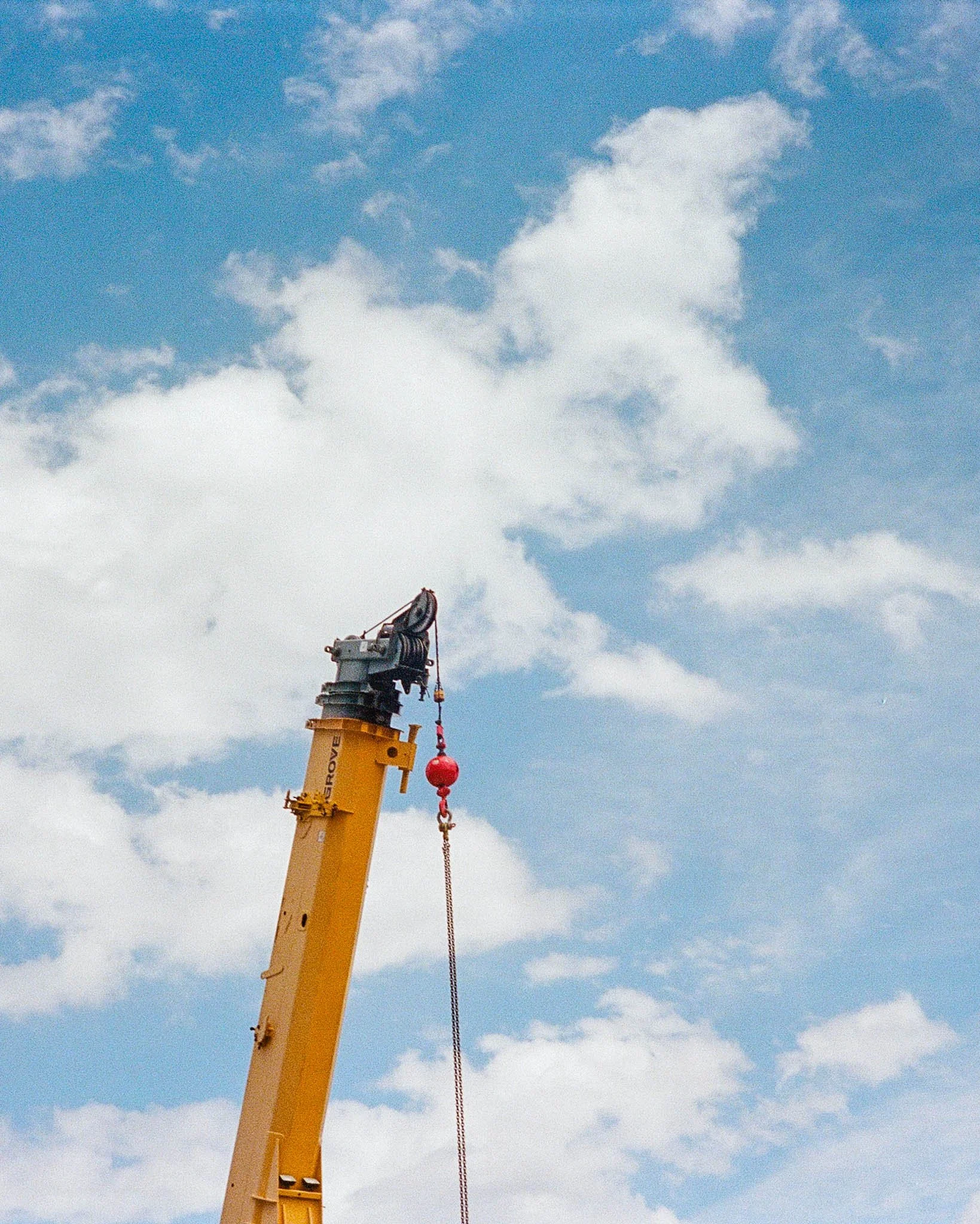 A yellow crane lifting a red ball on a chain with a cloudy blue sky in the background.