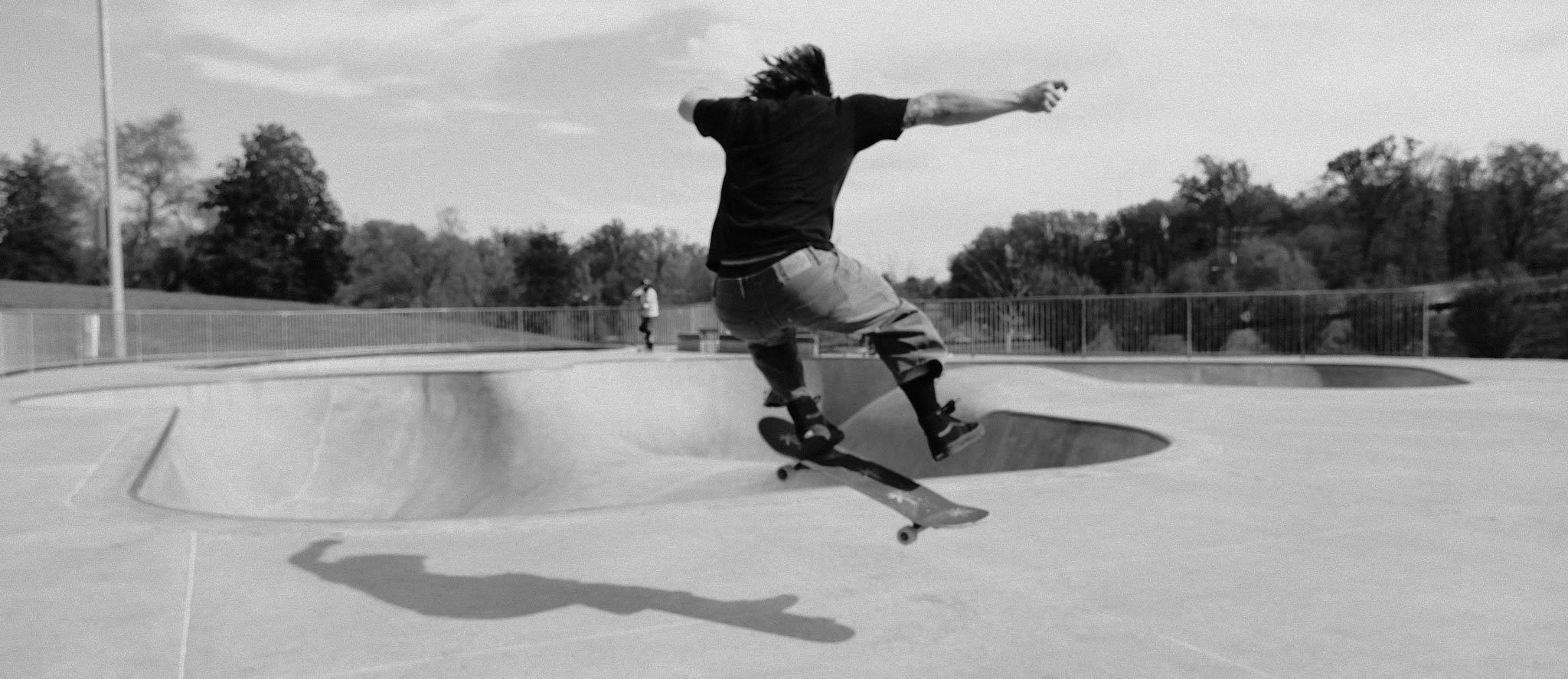 A skateboarder performing a trick at a skate park during the daytime, with trees and a fence in the background.
