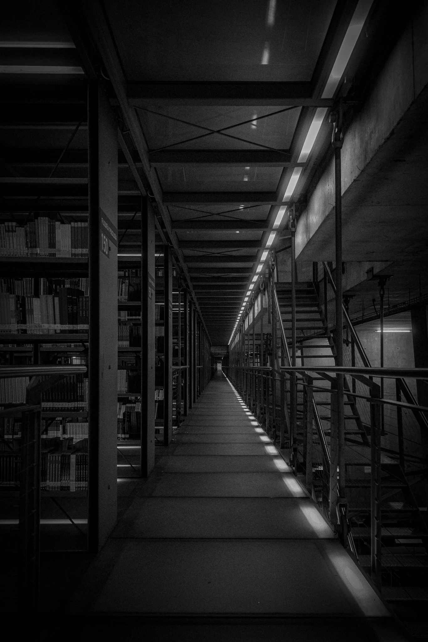 Black and white photo of a hallway with a metal railing on the right, bookshelves on the left, and stairs leading up on the right side, in a modern library or building.
