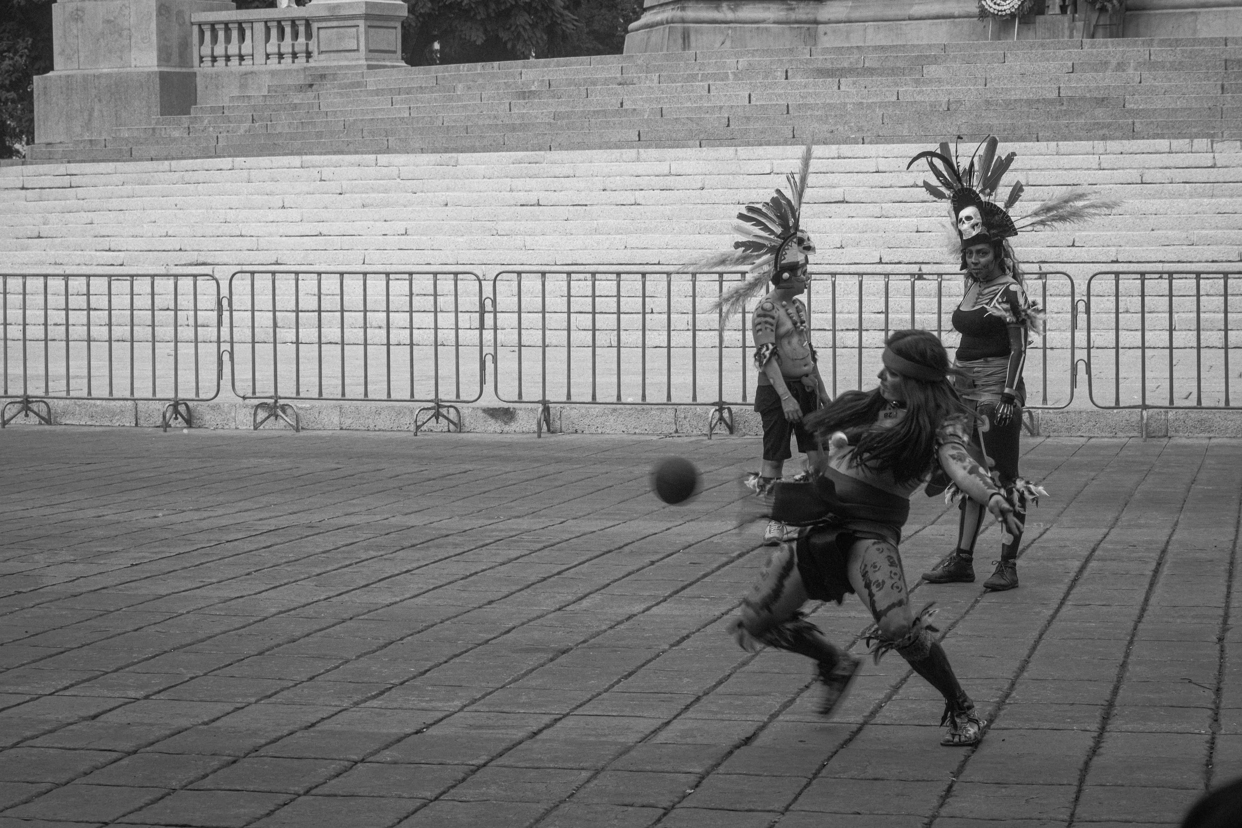 Group of performers in traditional costumes with feathers and skull accessories, playing with a ball on a paved open space, with steps and barriers in the background.