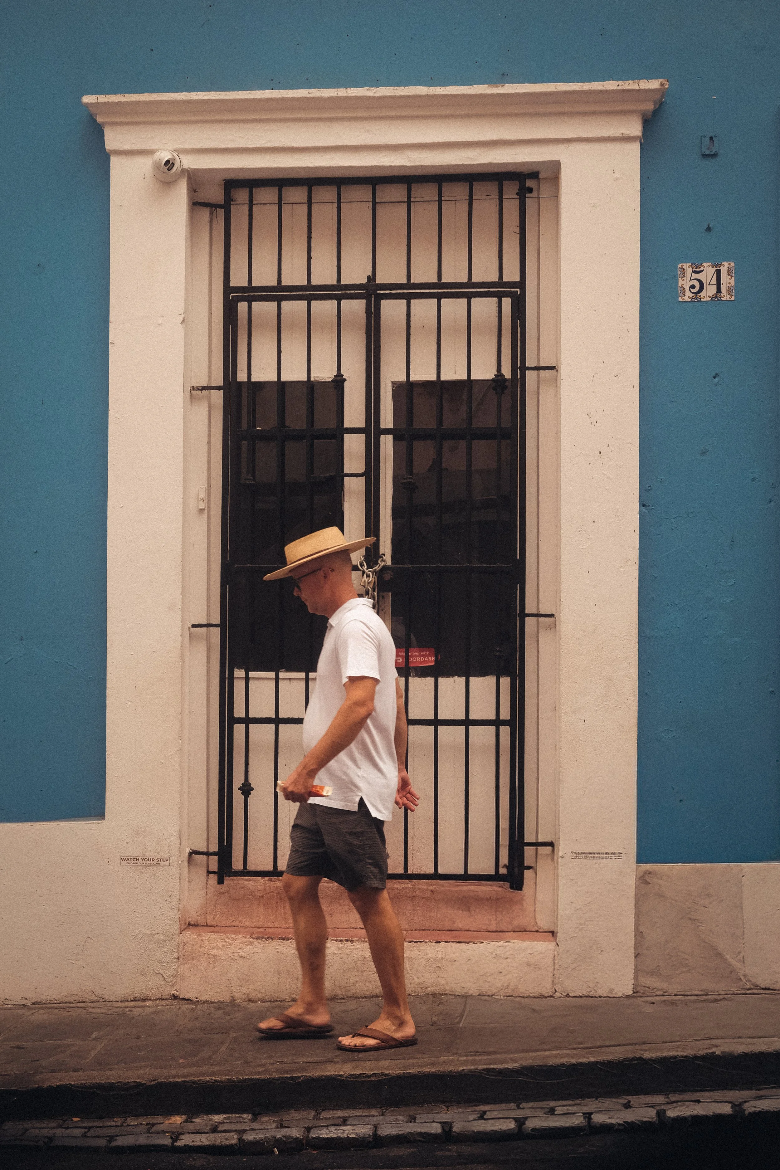 A man walking on a sidewalk in front of a colorful building with a blue and white facade, street number 51, wearing a broad-brimmed hat, white T-shirt, dark shorts, and flip-flops, holding a smartphone in his right hand.