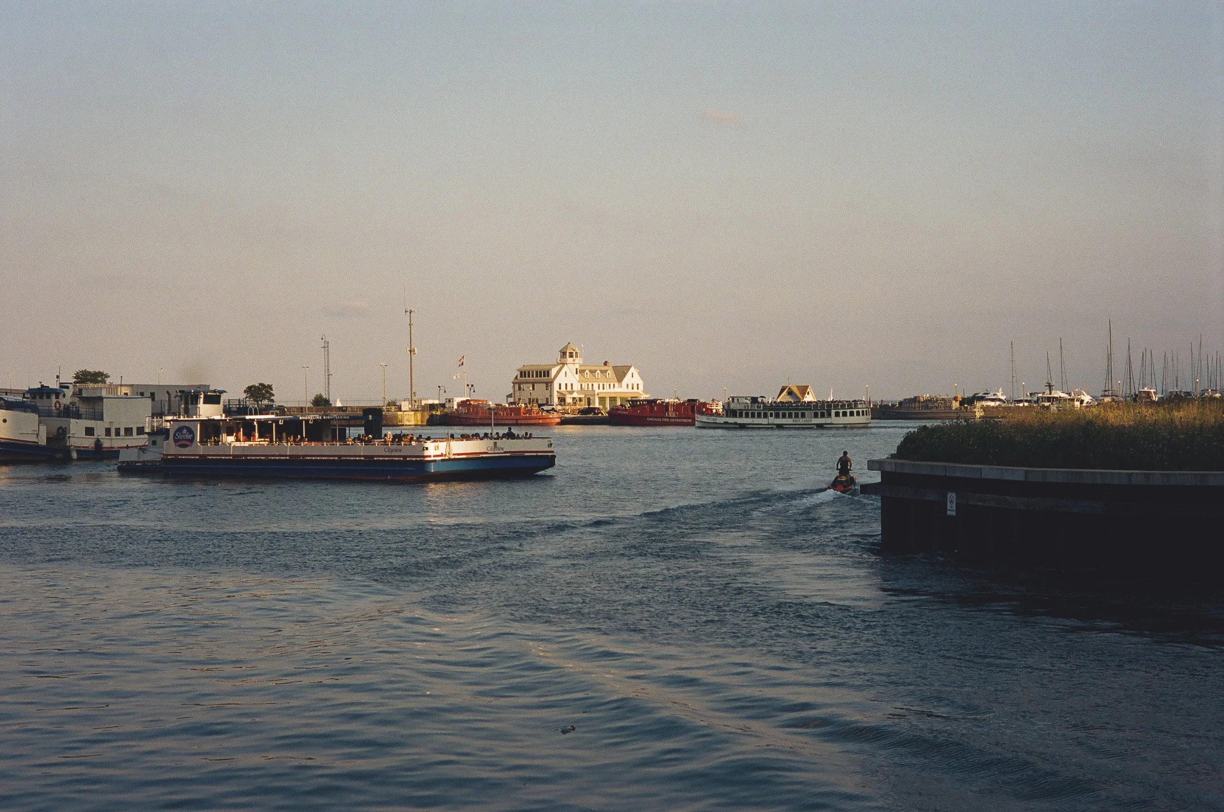 Boats and a small boat on a harbor with a lighthouse-style building in the background and a person riding a jet ski on the water.