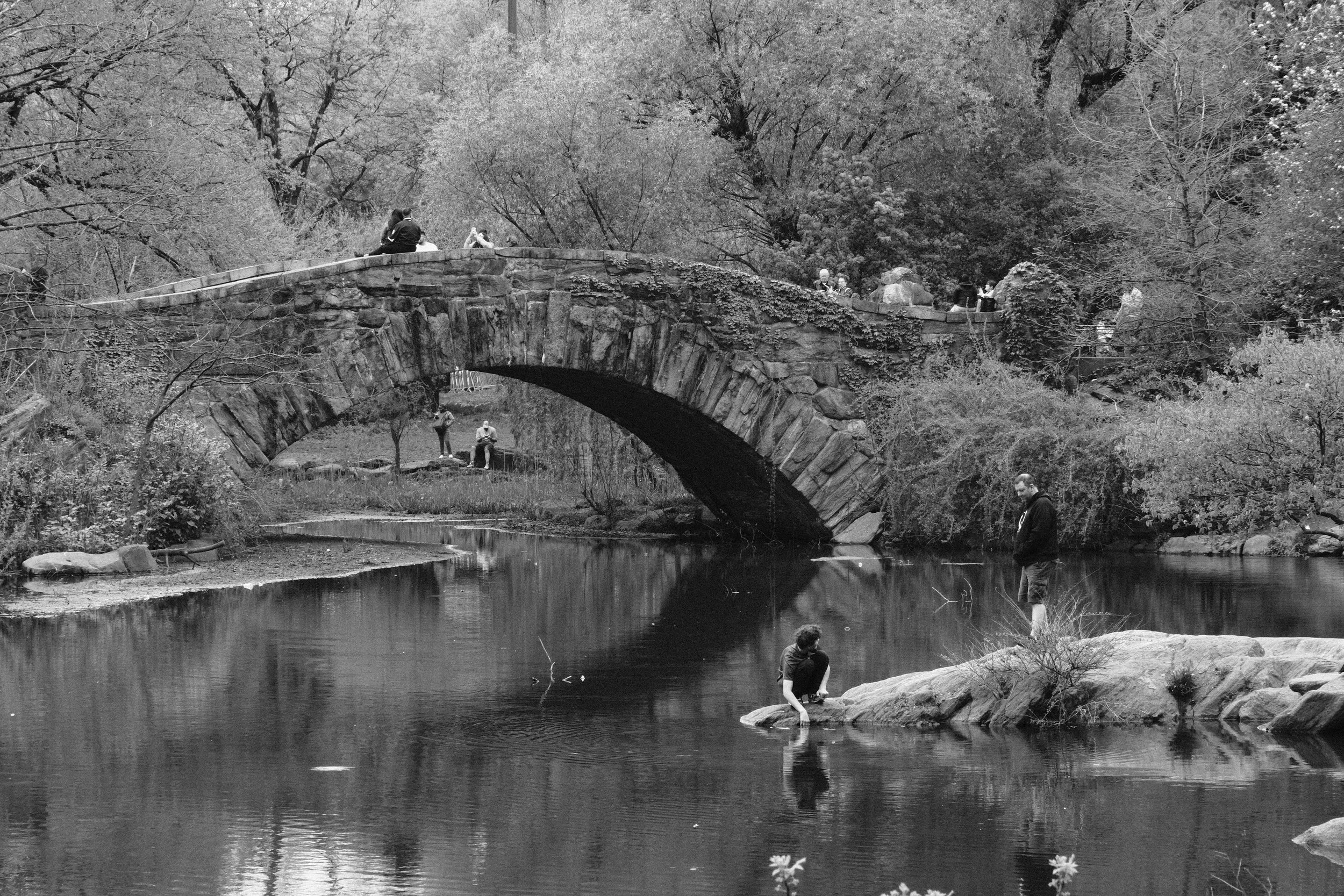 People gather around a stone arch bridge over a calm river in a wooded park, with some sitting on the bridge and others standing or sitting on rocks near the water.