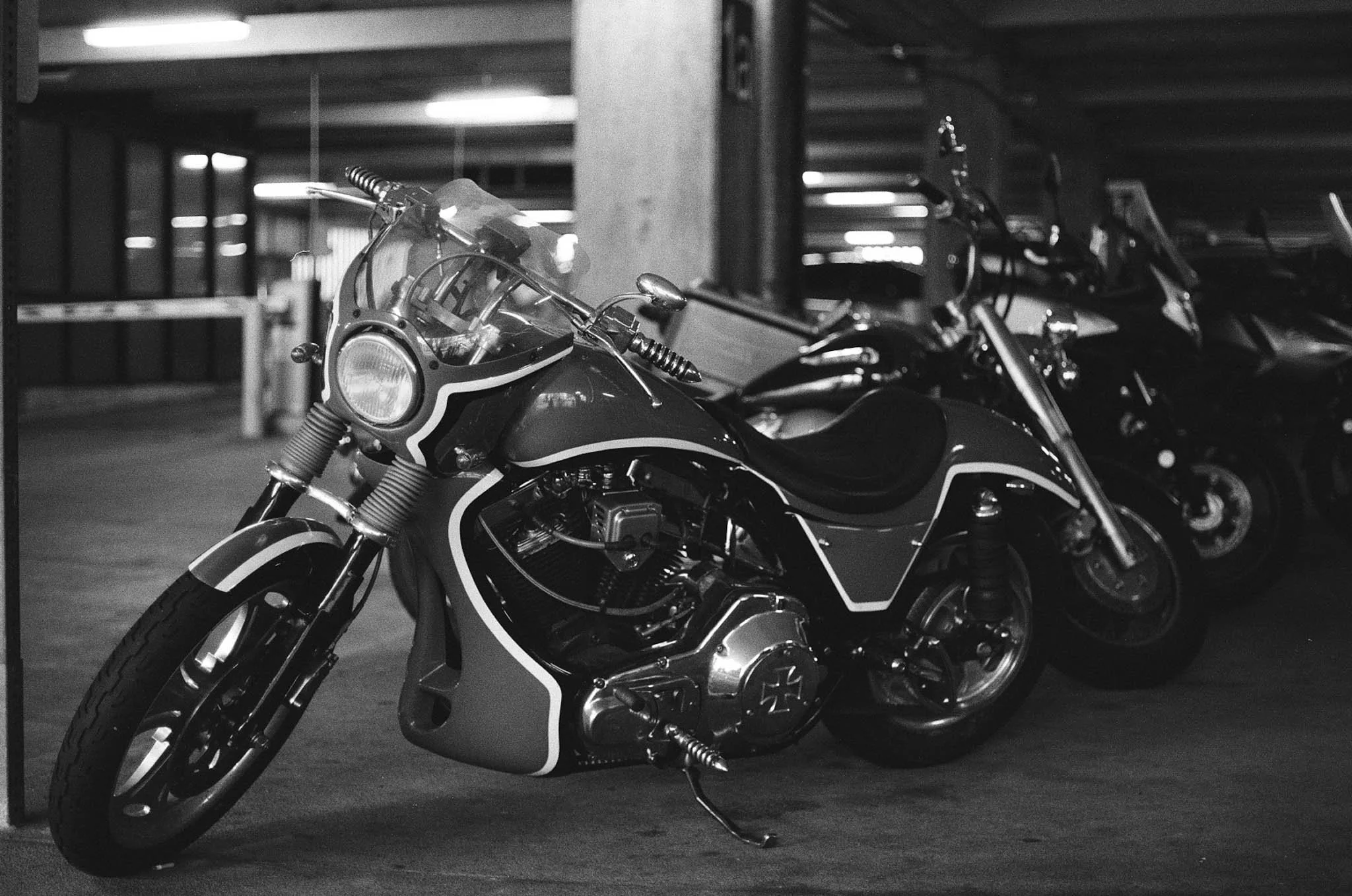 A black and white photograph of a motorcycle parked in an indoor parking garage, with other motorcycles lined up beside it.