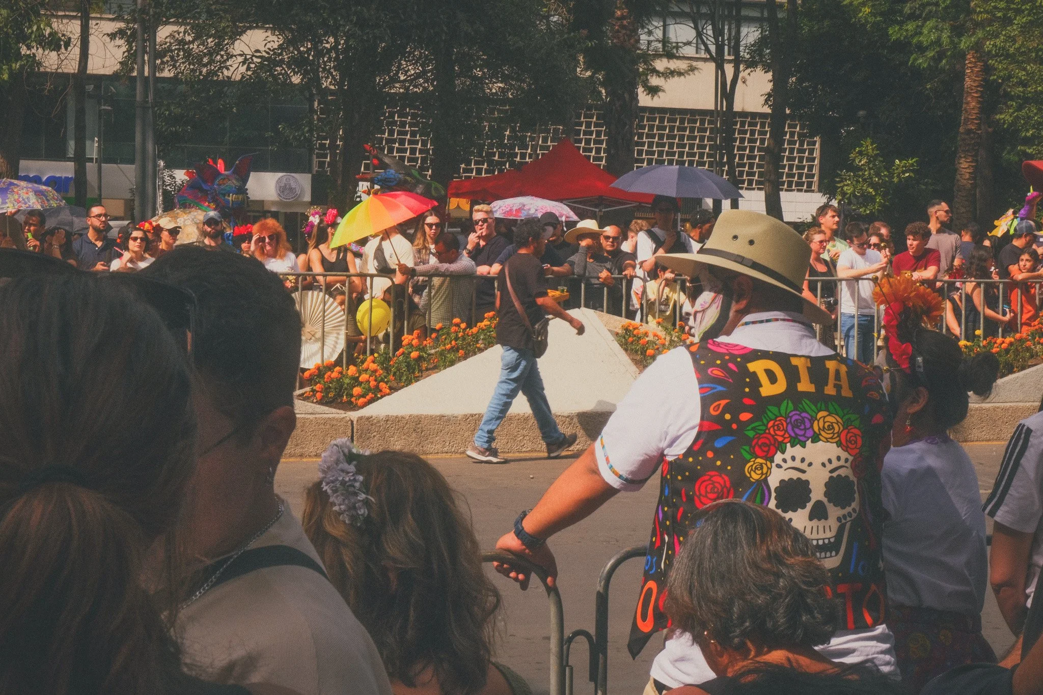 A lively outdoor event with a crowd of people behind metal barricades, some holding umbrellas. In the foreground, there are a few people including a person wearing a colorful vest with a skull and flower design, and another person with a floral acces