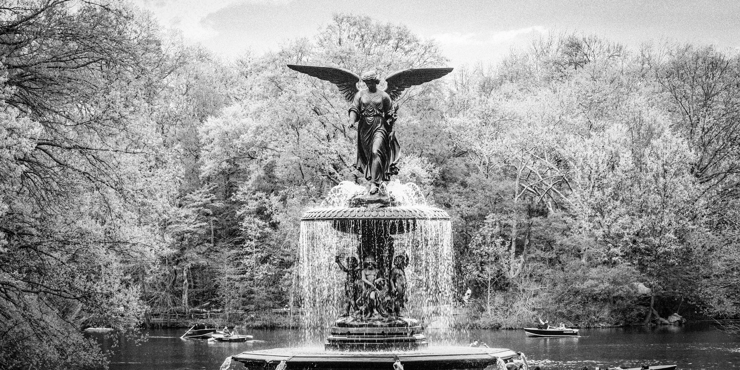A black-and-white photo of a large ornate fountain in a park with a statue of an angel with wings at the top, water cascading from the fountain, and people in boats on the water.