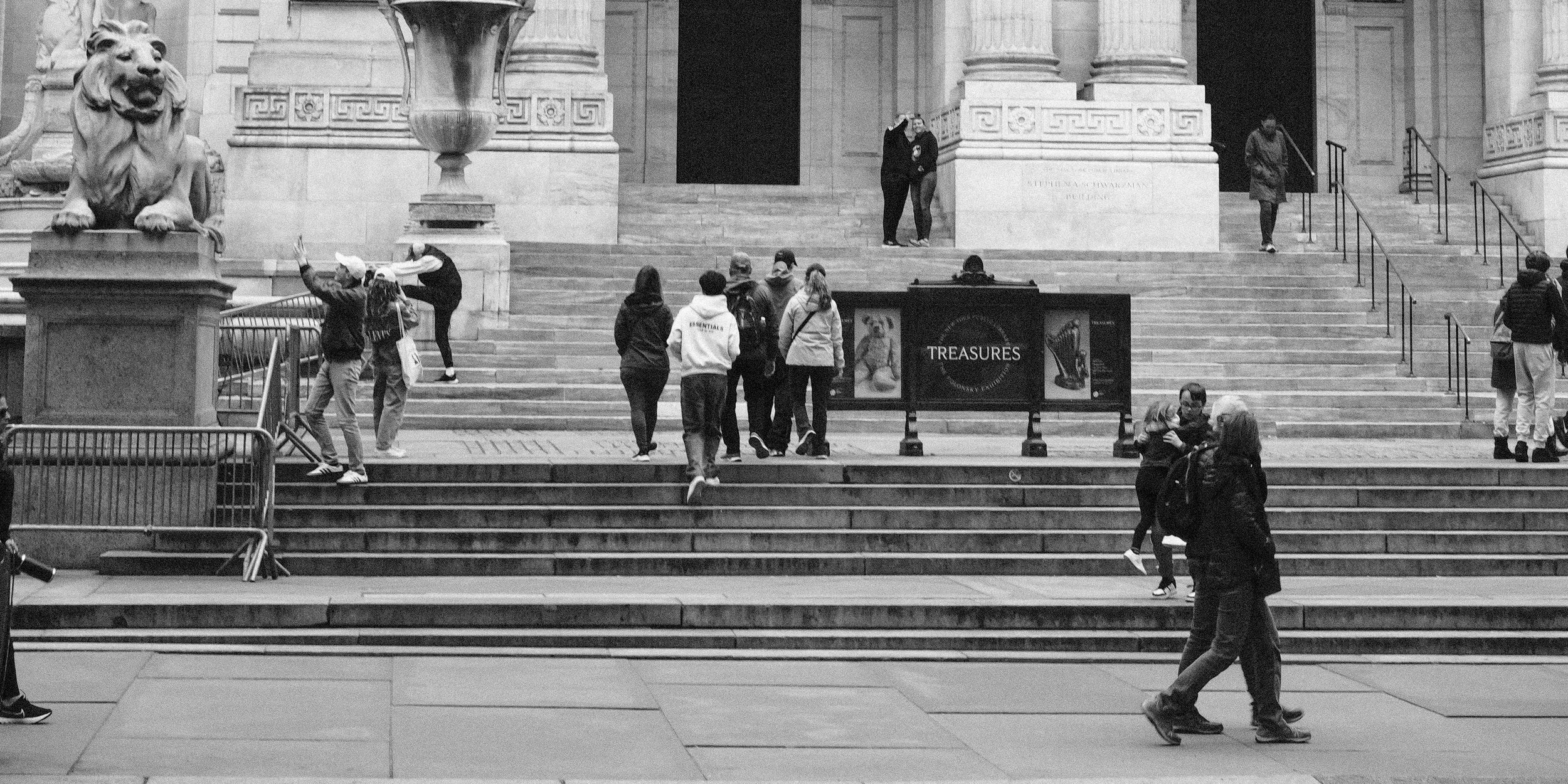 People walking and gathering on the steps of a historical building with large columns and lion statues at either side.