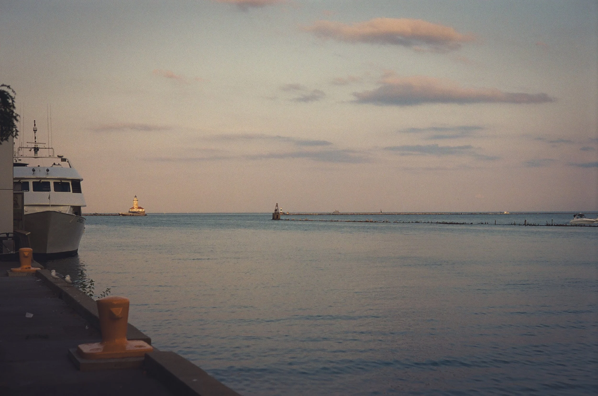 A harbor scene at dusk with a large white yacht docked on the left, a lighthouse in the distance on the left, and a breakwater extending into the water with a small lighthouse at the end. The sky is partly cloudy with a soft glow, reflecting on the c