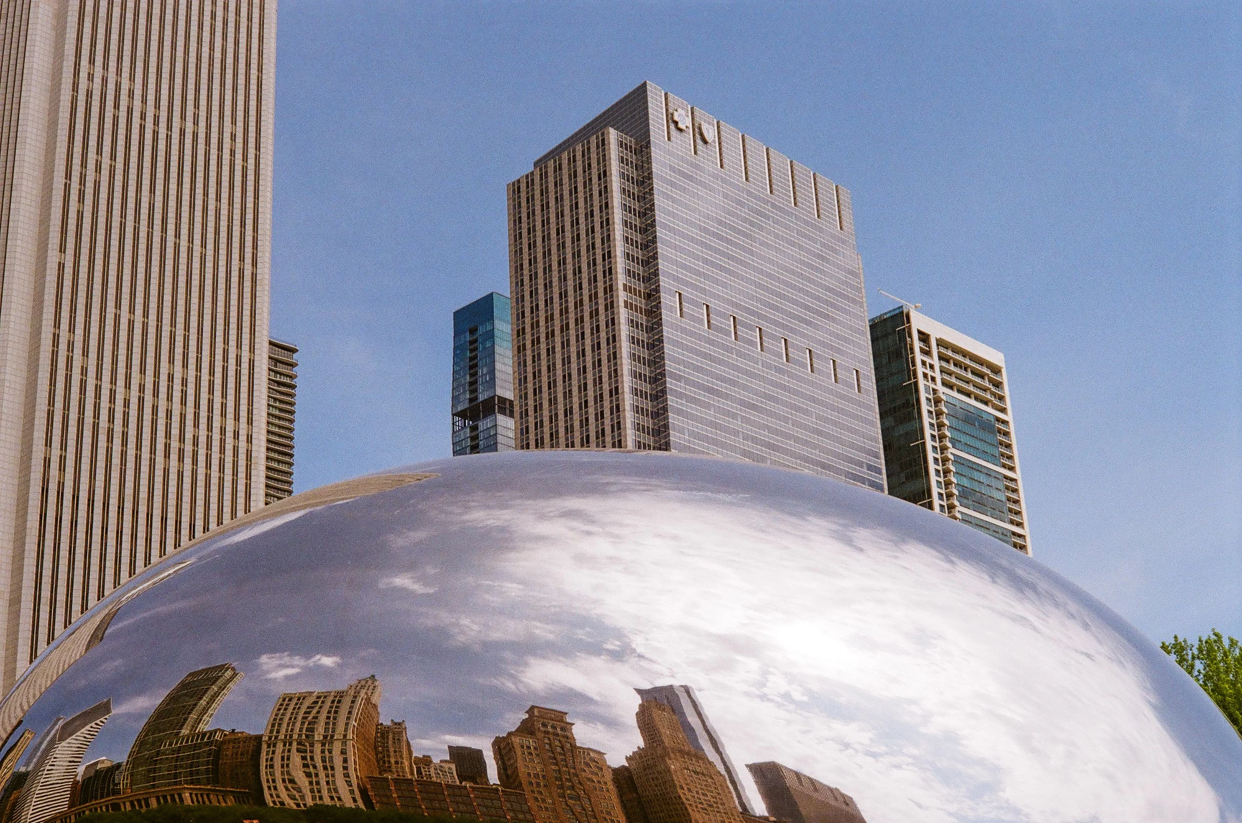 Reflected city skyline in a large, shiny, metallic sculpture with tall modern buildings and a blue sky.