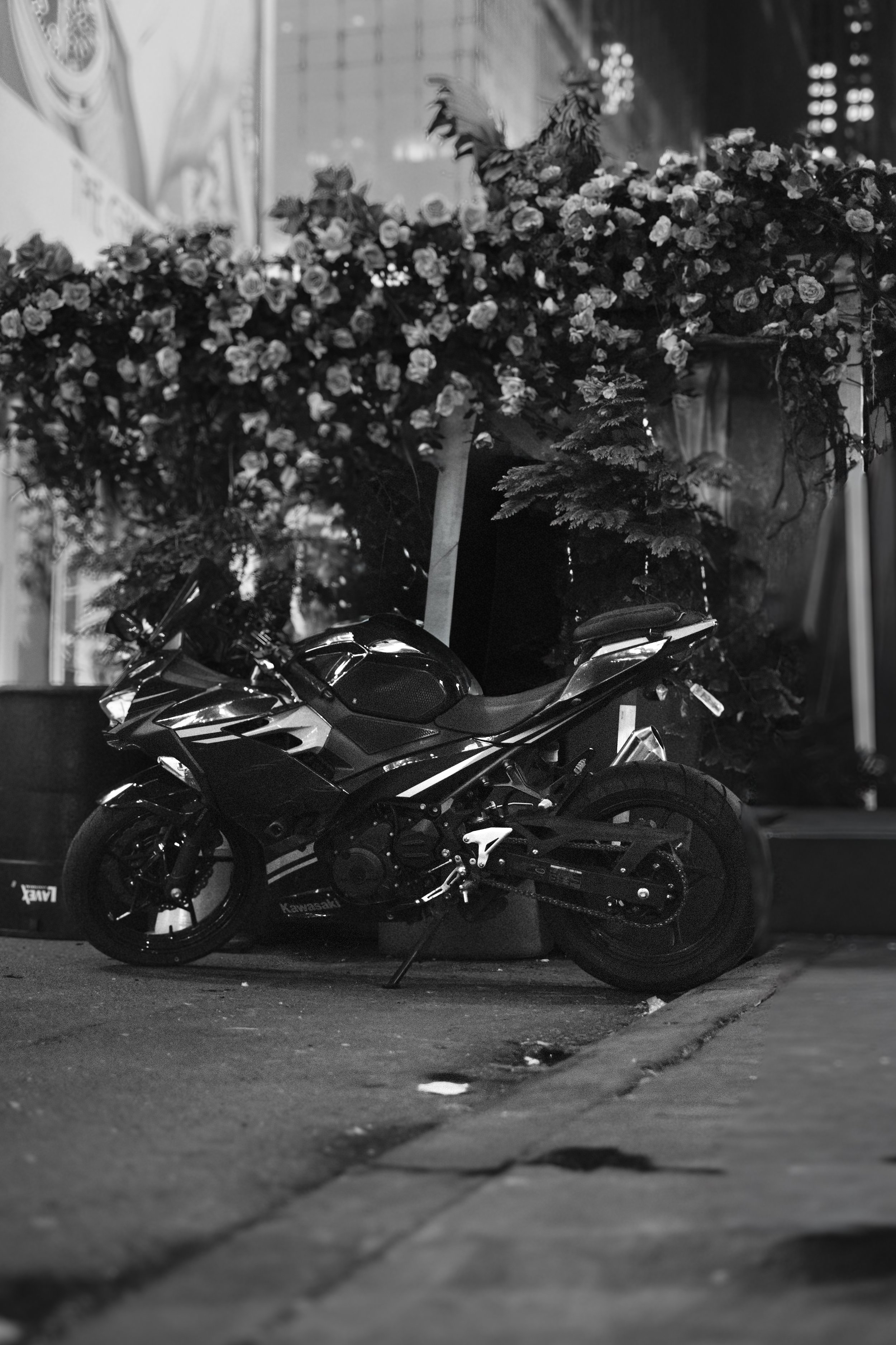 A black motorcycle parked on the sidewalk in front of a floral display and trees, captured at night in black and white.