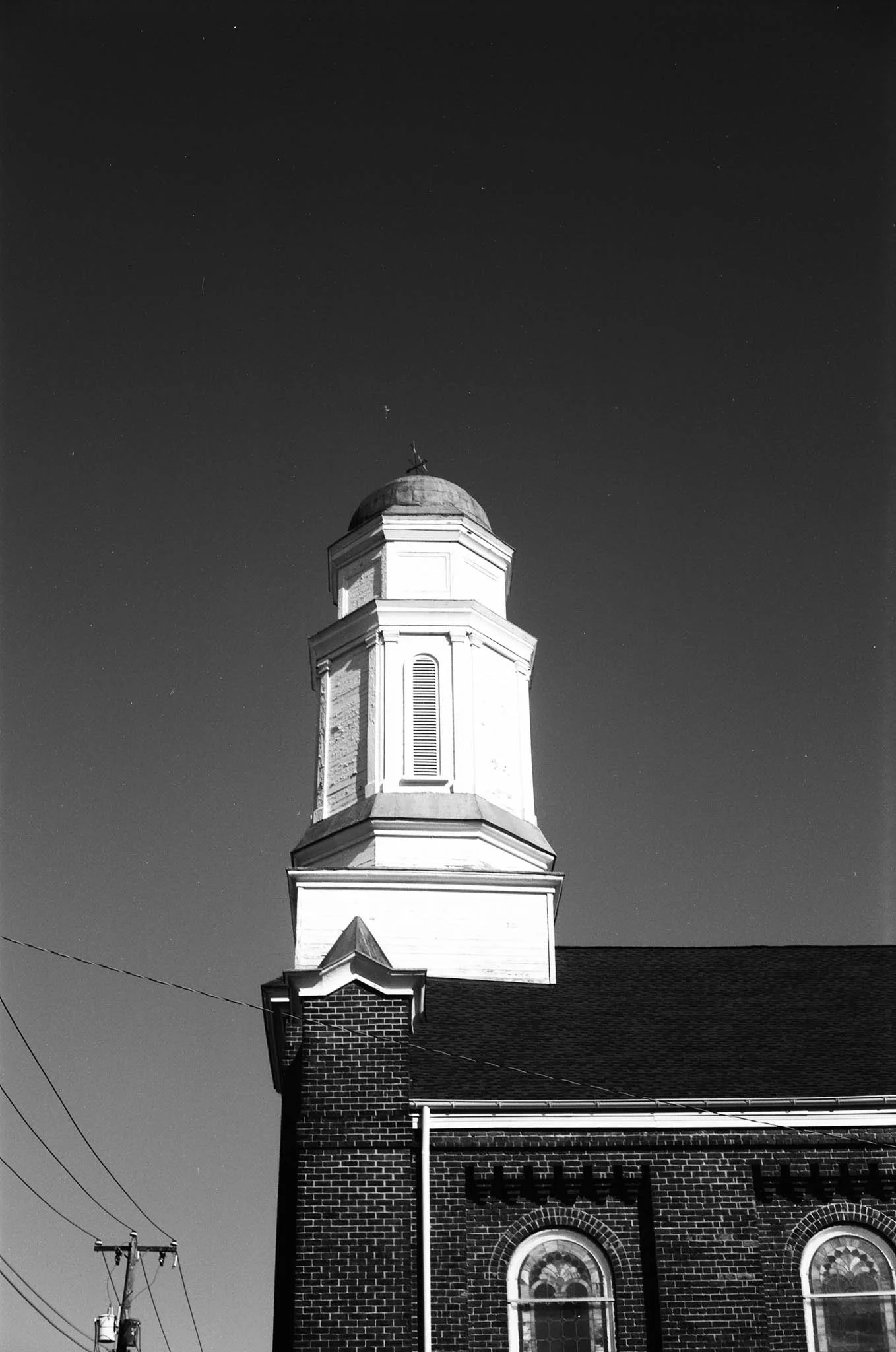 Black and white photograph of a church steeple with a cross on top, part of a brick church building with arched windows, against a clear sky.