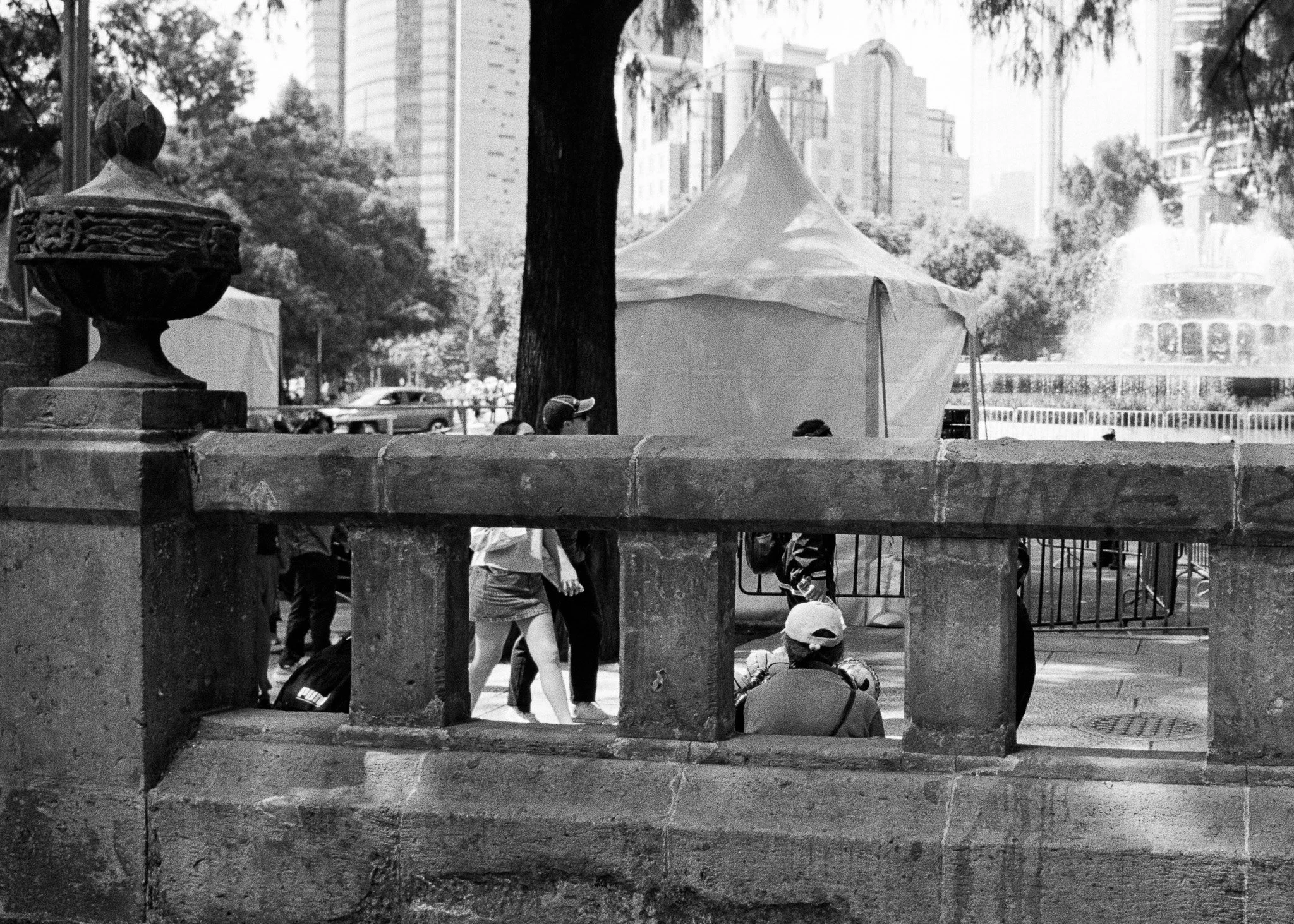 Black and white photo of a park scene with a stone wall in the foreground. Behind the wall, a person sits on the ground wearing a cap and a backpack, with their back to the camera. Several other people are walking or standing nearby. There is a large