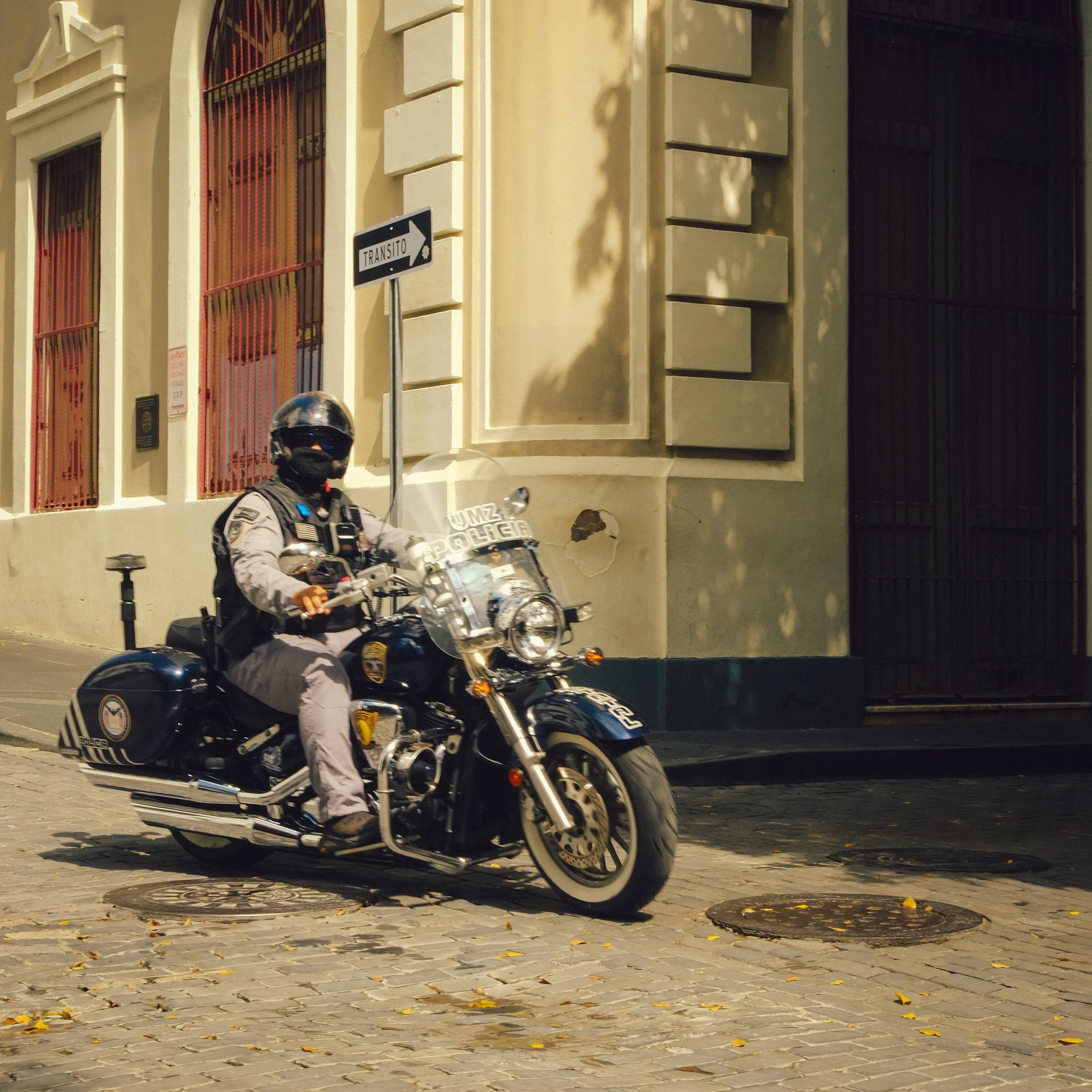 A police officer riding a black motorcycle on a cobblestone street next to a beige building with architectural details and red window bars.