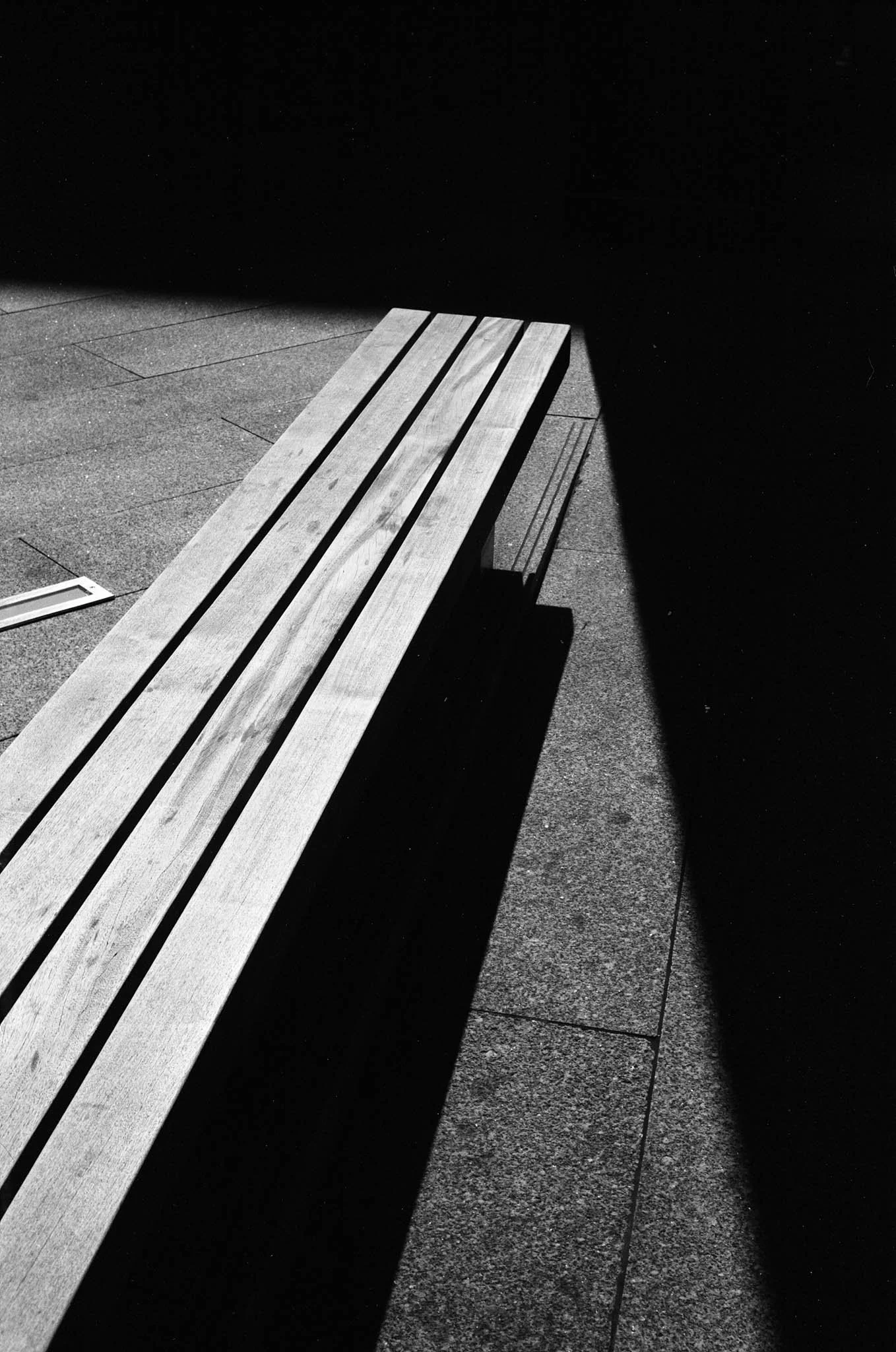 A wooden bench casting a shadow on a concrete sidewalk, with sunlight creating a stark contrast between light and dark areas.