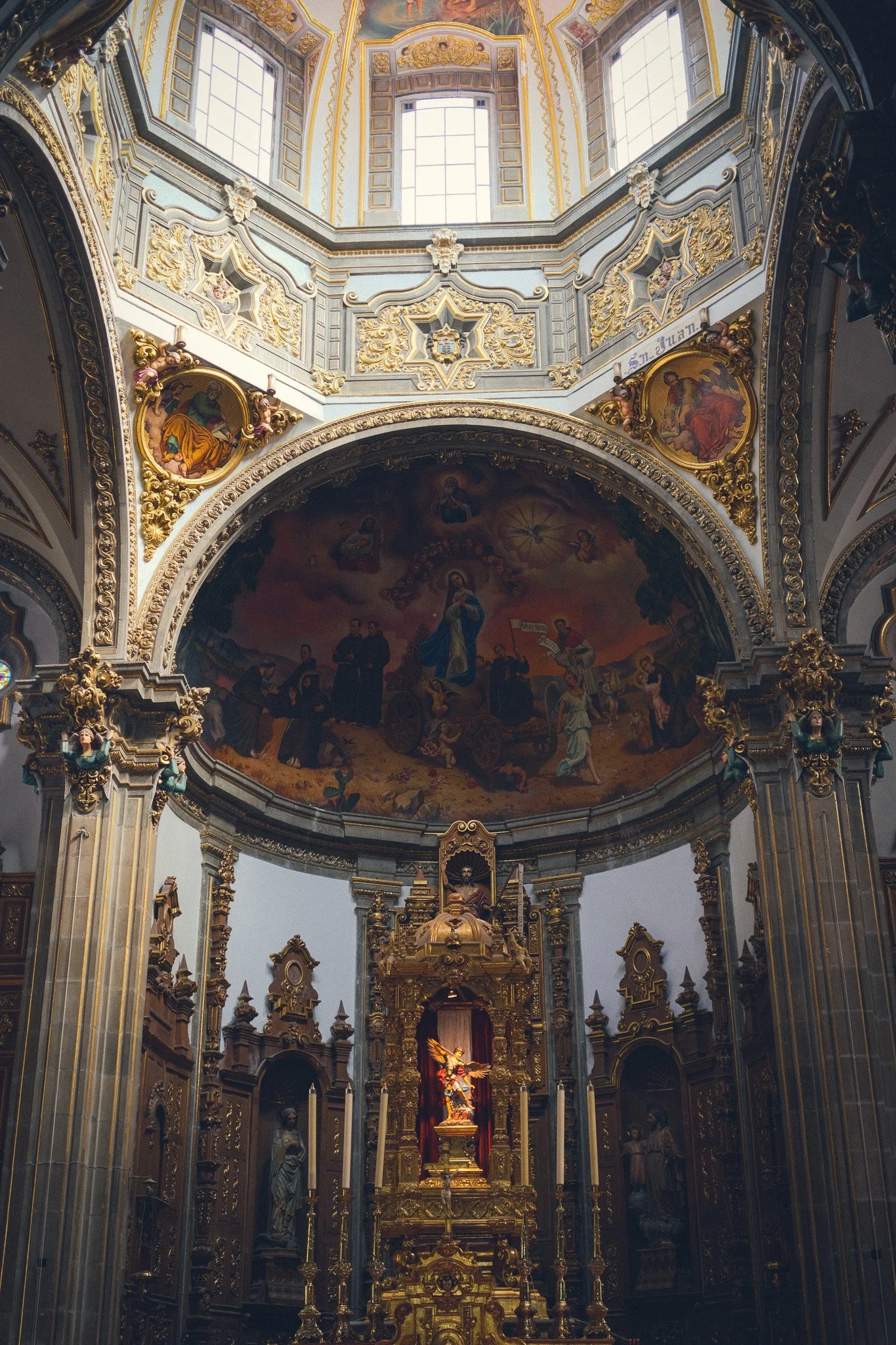 Interior of a church featuring ornate gold and wood detailing, a large mural on the ceiling depicting religious figures, and an altar with a statue of Jesus surrounded by candles.