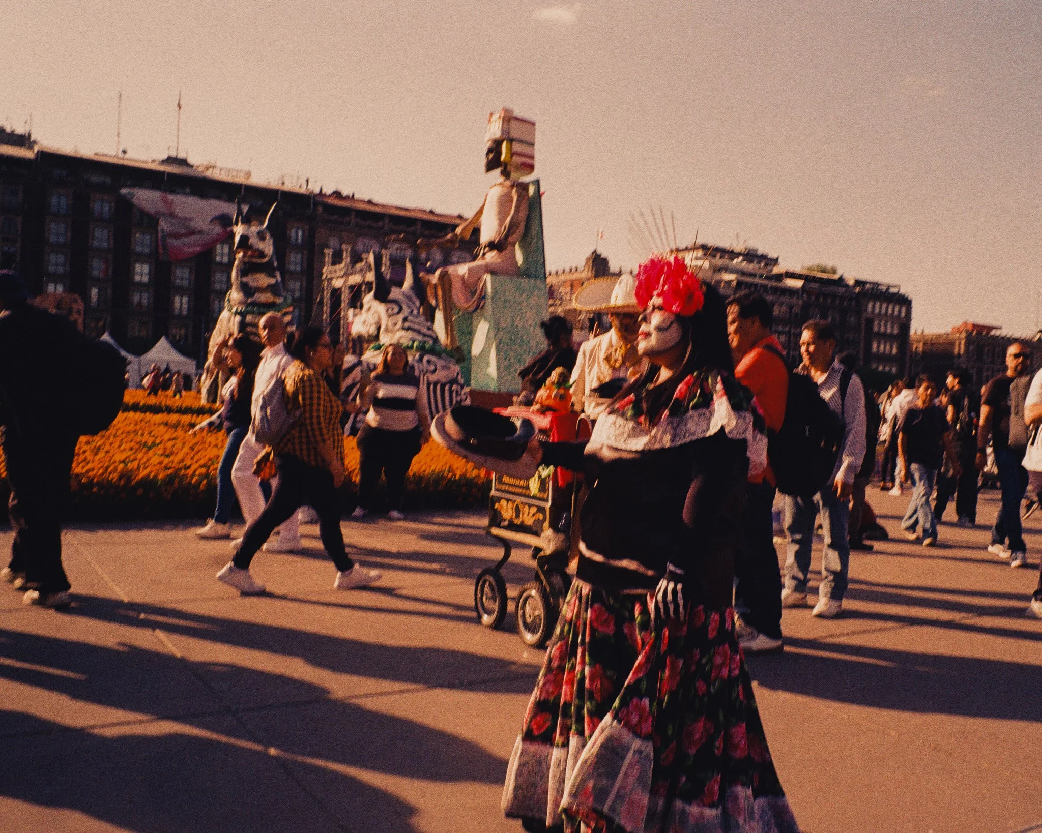 A street performer dressed as a traditional Mexican Catrina with face paint and a large red floral headpiece, holding a tray in front of a crowd walking in a festive outdoor event, with a float decorated with a skeleton figure in the background.