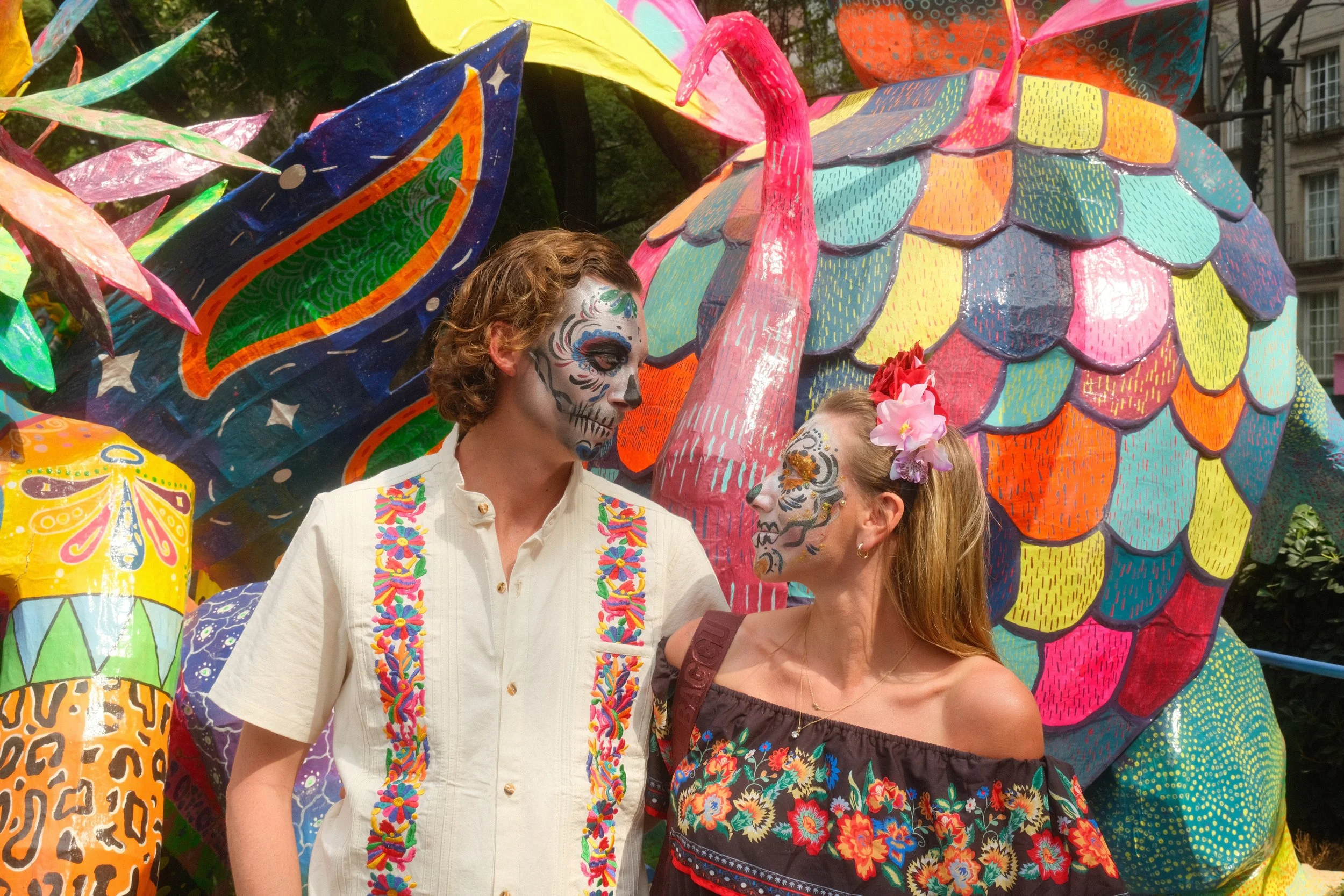Two people with face paint resembling Día de Muertos skulls, standing close together in front of colorful, large paper-mâché sculptures of animals and abstract shapes at a festival.