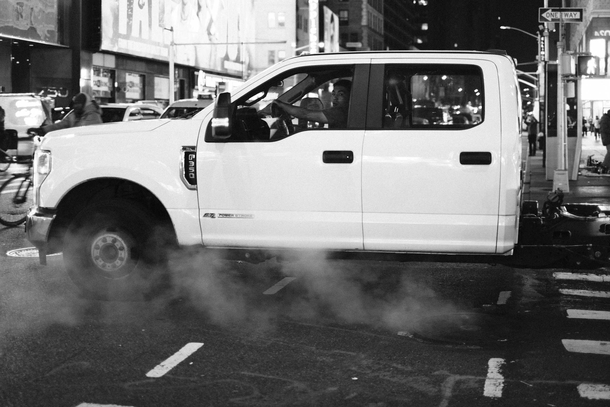A white Ford F350 pickup truck on a city street at night with steam rising from the pavement, seen in black and white. People and neon lights are visible in the background.