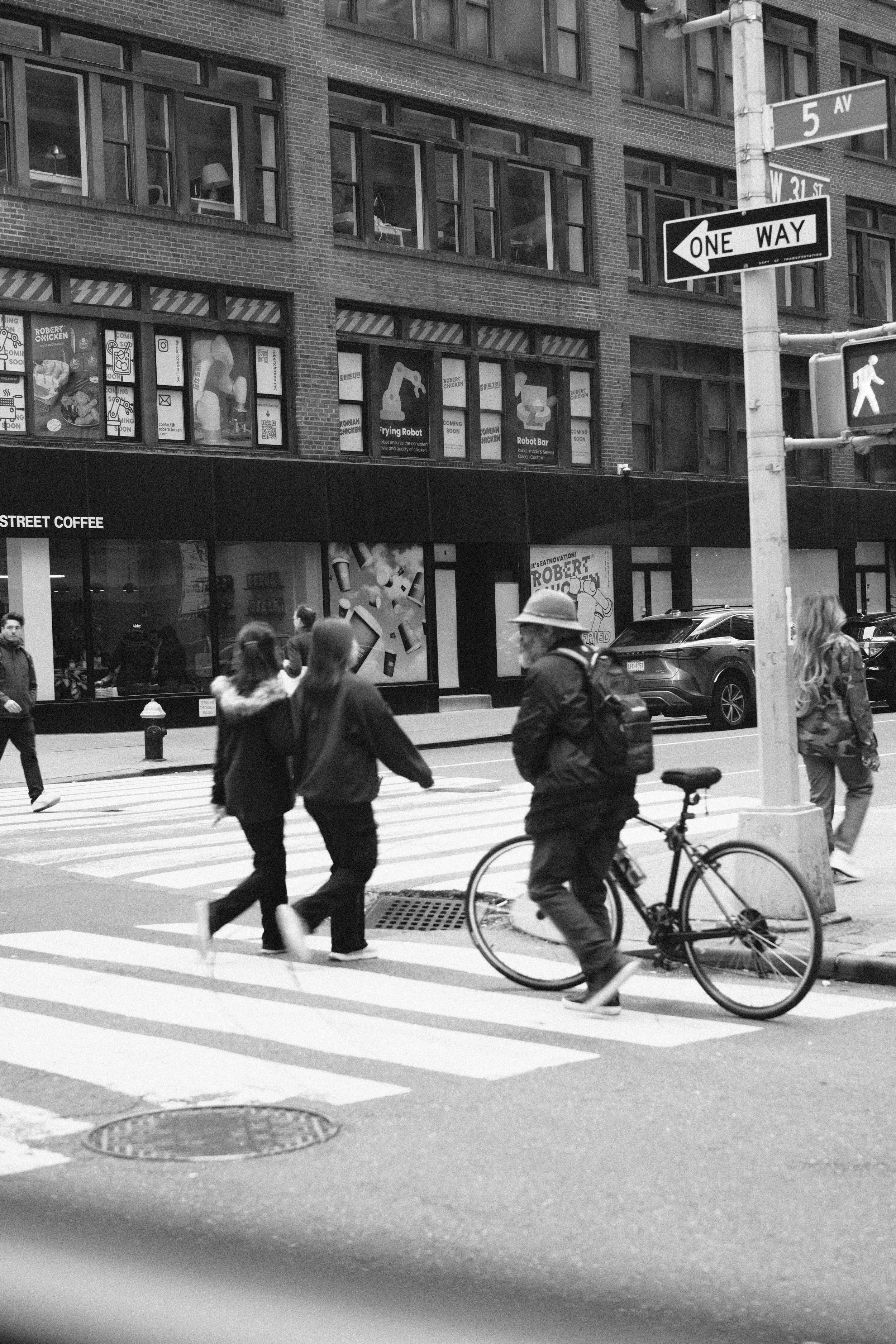Black and white photo of pedestrians crossing the street on a city crosswalk. A cyclist with a backpack and helmet is waiting at the corner. There are buildings with large windows and signage on the shops, including a coffee shop and a store advertis