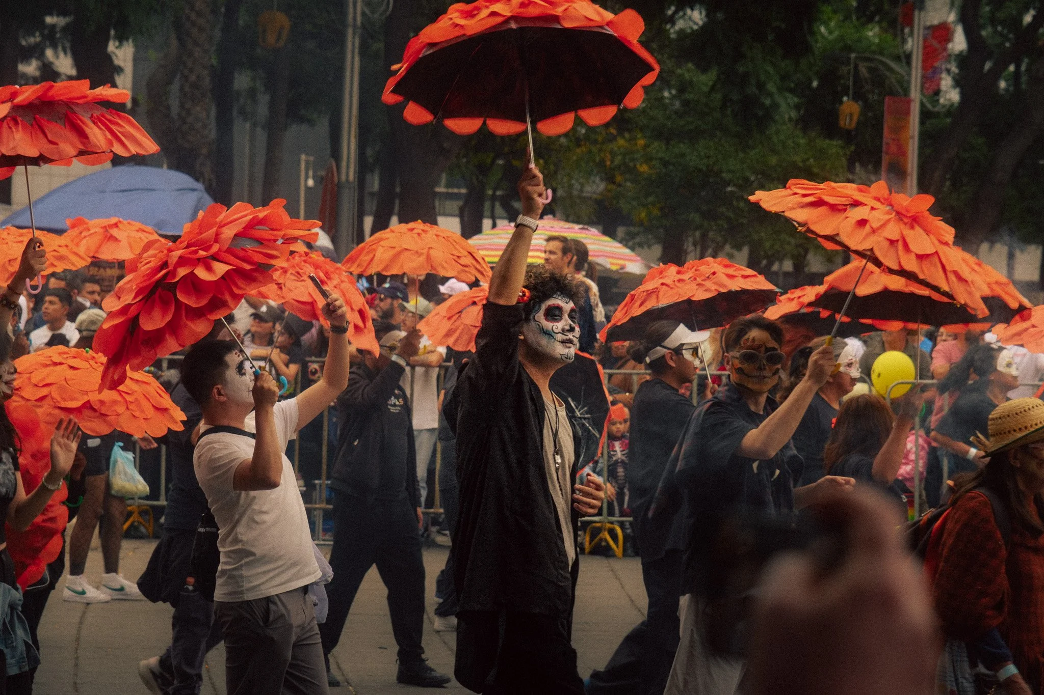 People participating in a parade wearing skull makeup and holding orange umbrellas, celebrating Day of the Dead.