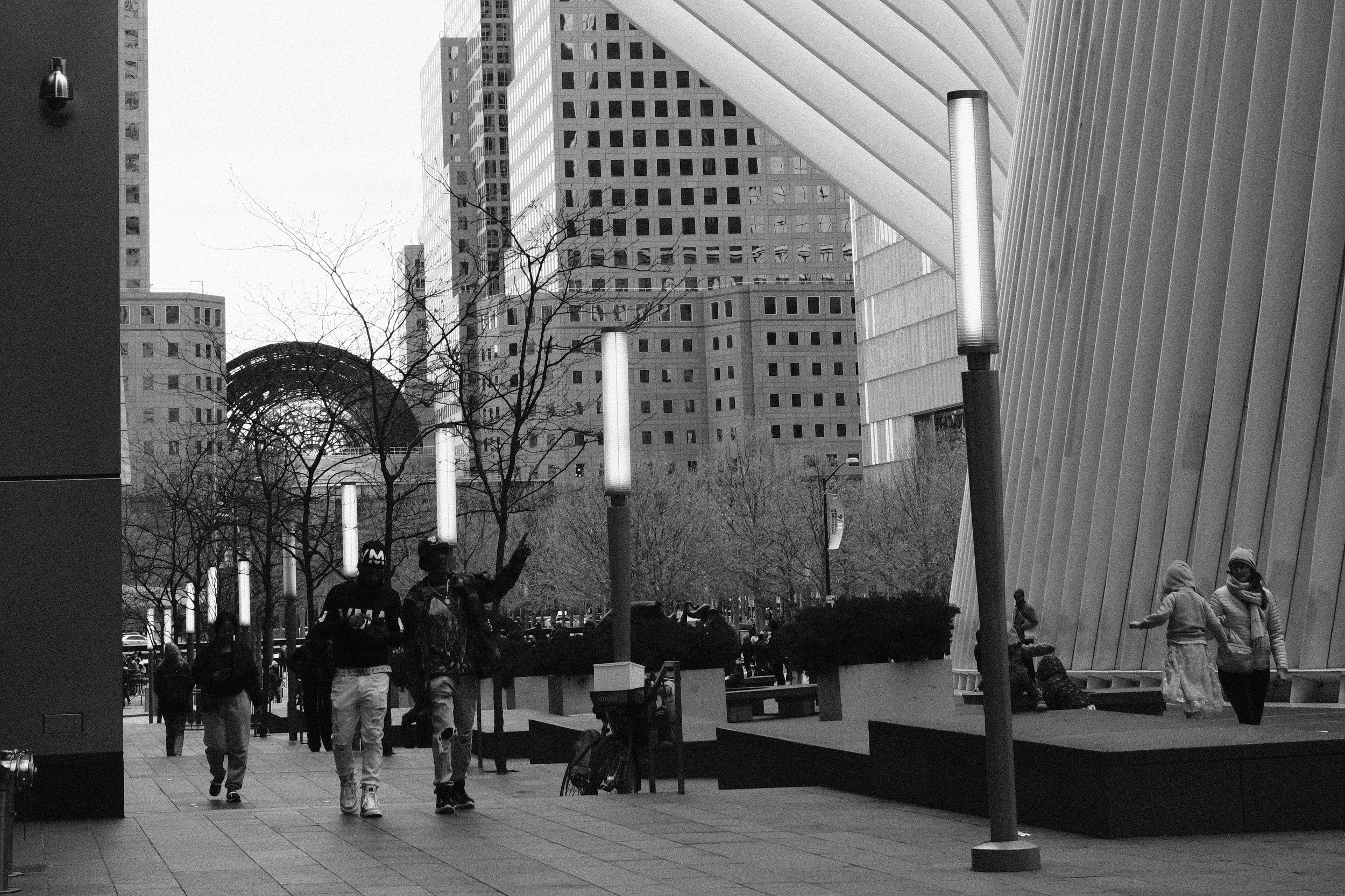 Black and white photo of people walking in an urban park area with modern buildings in the background, featuring trees, benches, and street lamps.
