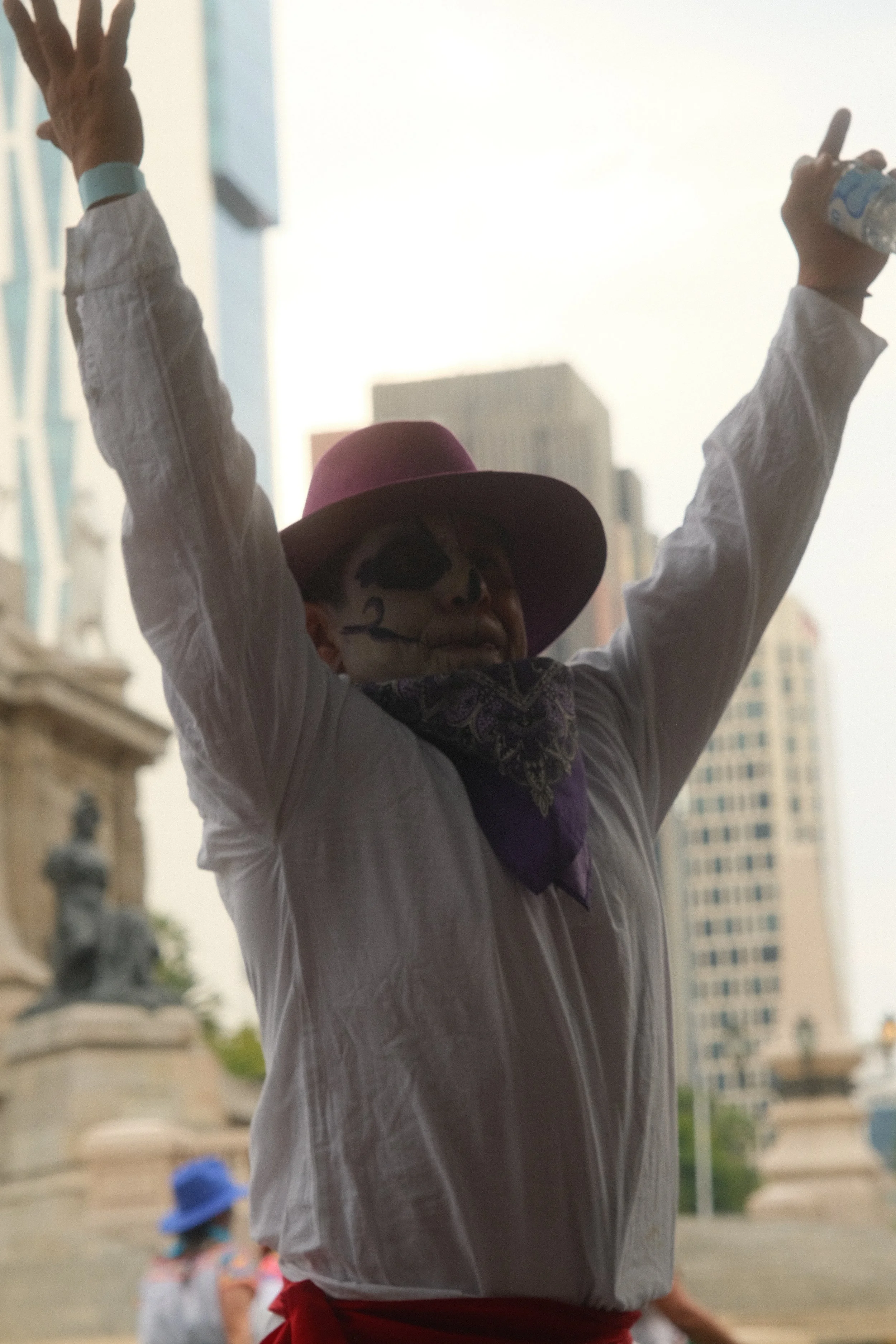 Person with face painted as a skeleton, wearing a wide-brimmed purple hat and bandana, raising arms and holding a water bottle in an urban park setting.
