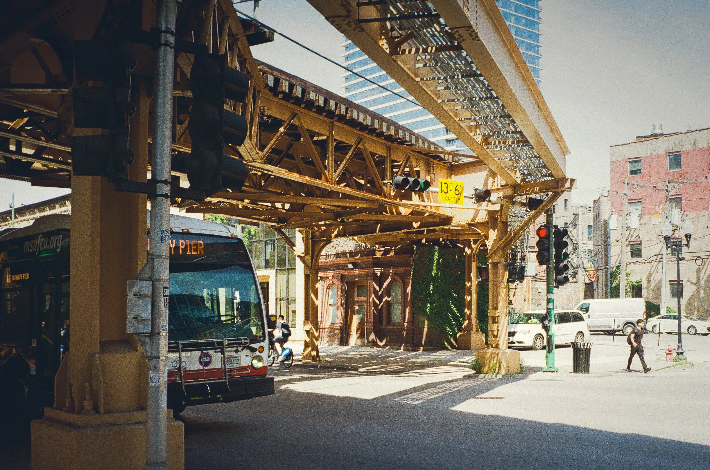An urban scene with a bus at a bus stop on a city street, elevated train tracks overhead, traffic lights, pedestrians, and background buildings.