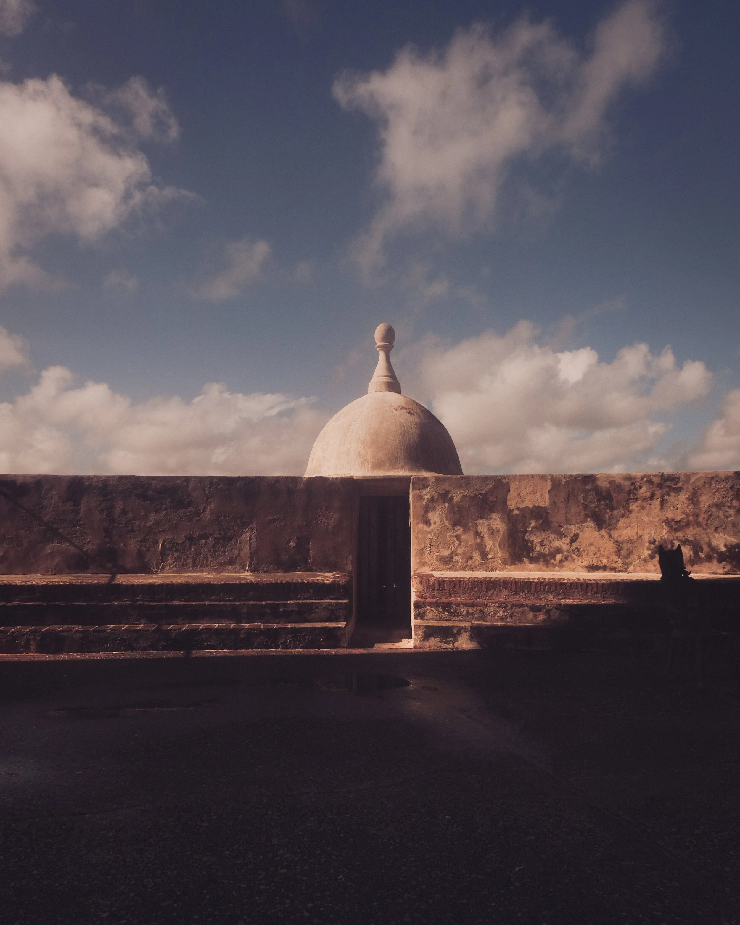 A tall, white domed structure with a finial sits behind a stone wall with stairs leading up to it. The sky is partly cloudy with sunlight casting shadows on the wall. A black cat is sitting on the right side of the wall.