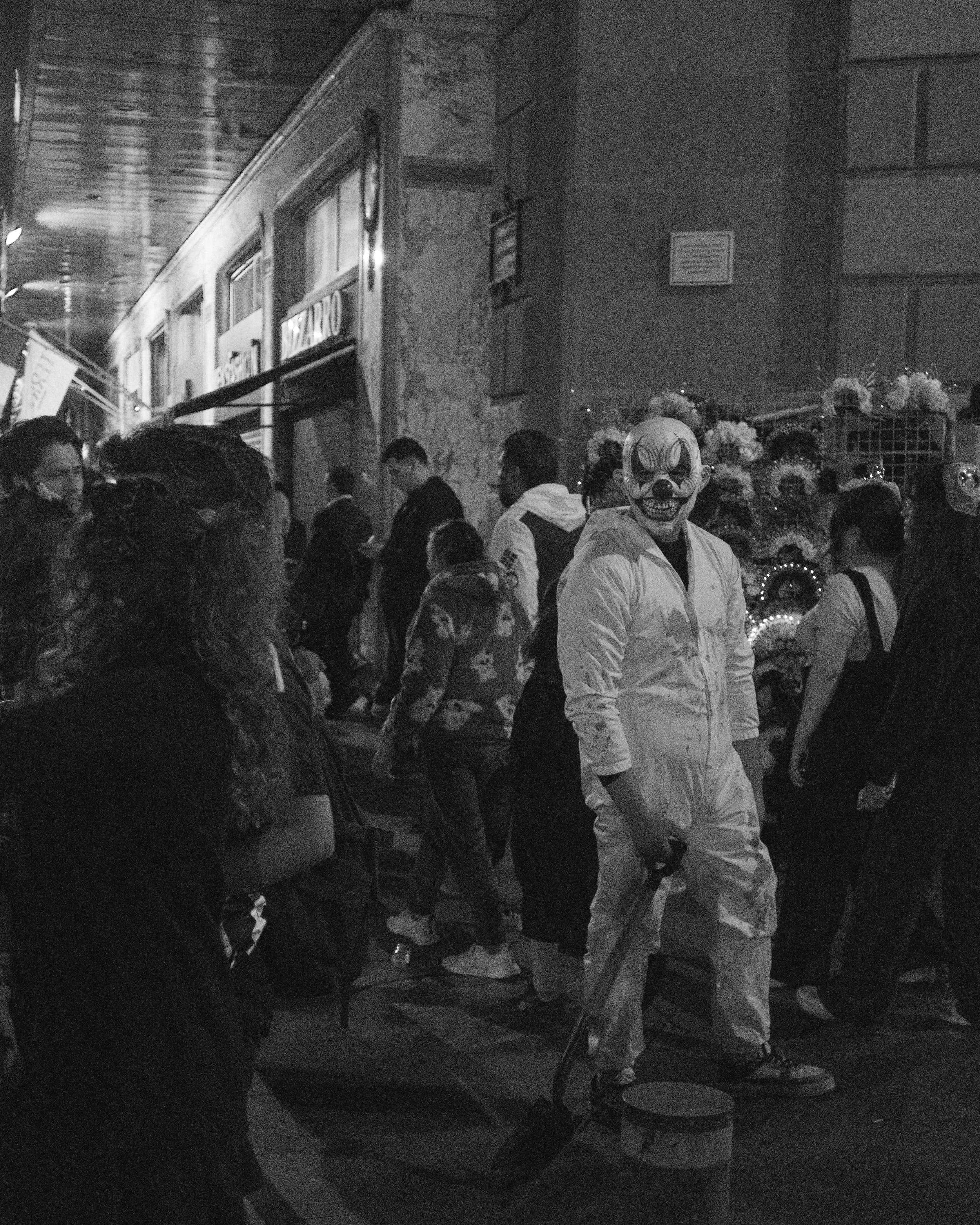A person dressed in a scary clown mask and white coveralls stands in a crowded outdoor street scene at night. The background includes people, a store with flower decorations, and a lit-up sign.
