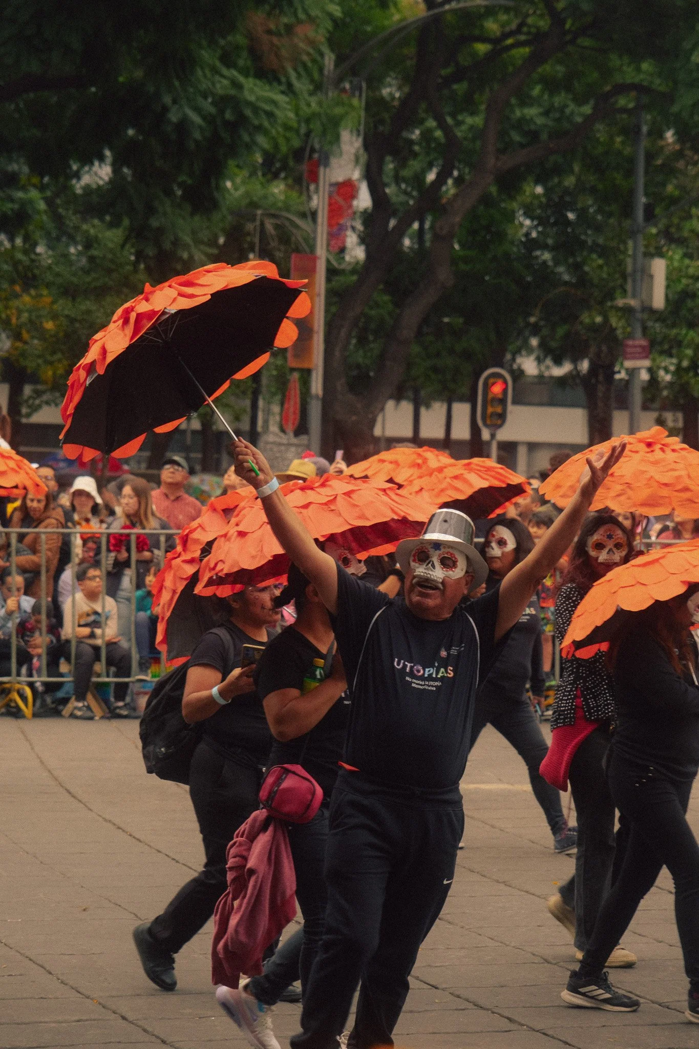 People participating in a parade wearing skeleton masks and holding orange umbrellas, with a crowd watching behind a barricade in a city street.