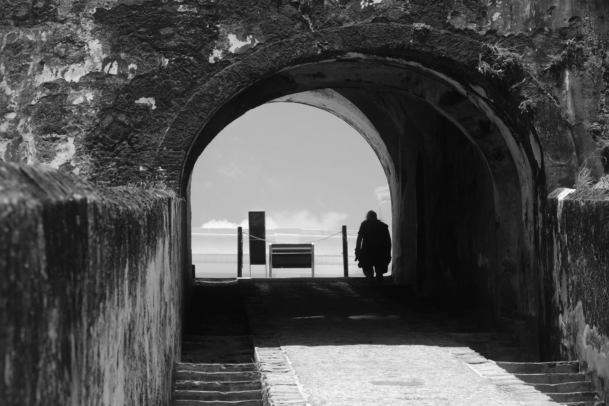 A person walking through a tunnel with stone walls, headed toward an open sky and a bench outside.