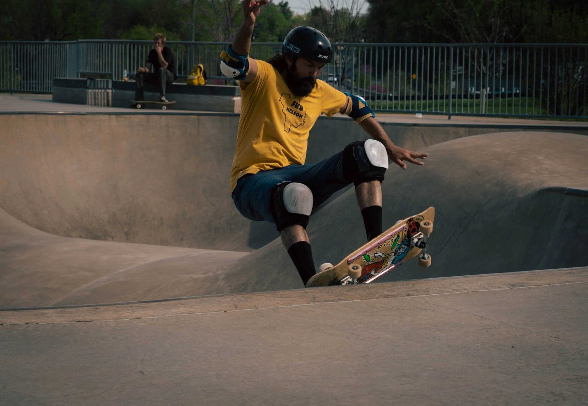 Skateboarder in a skatepark wearing a helmet, yellow shirt, knee and elbow pads, performing a trick on a skateboard.