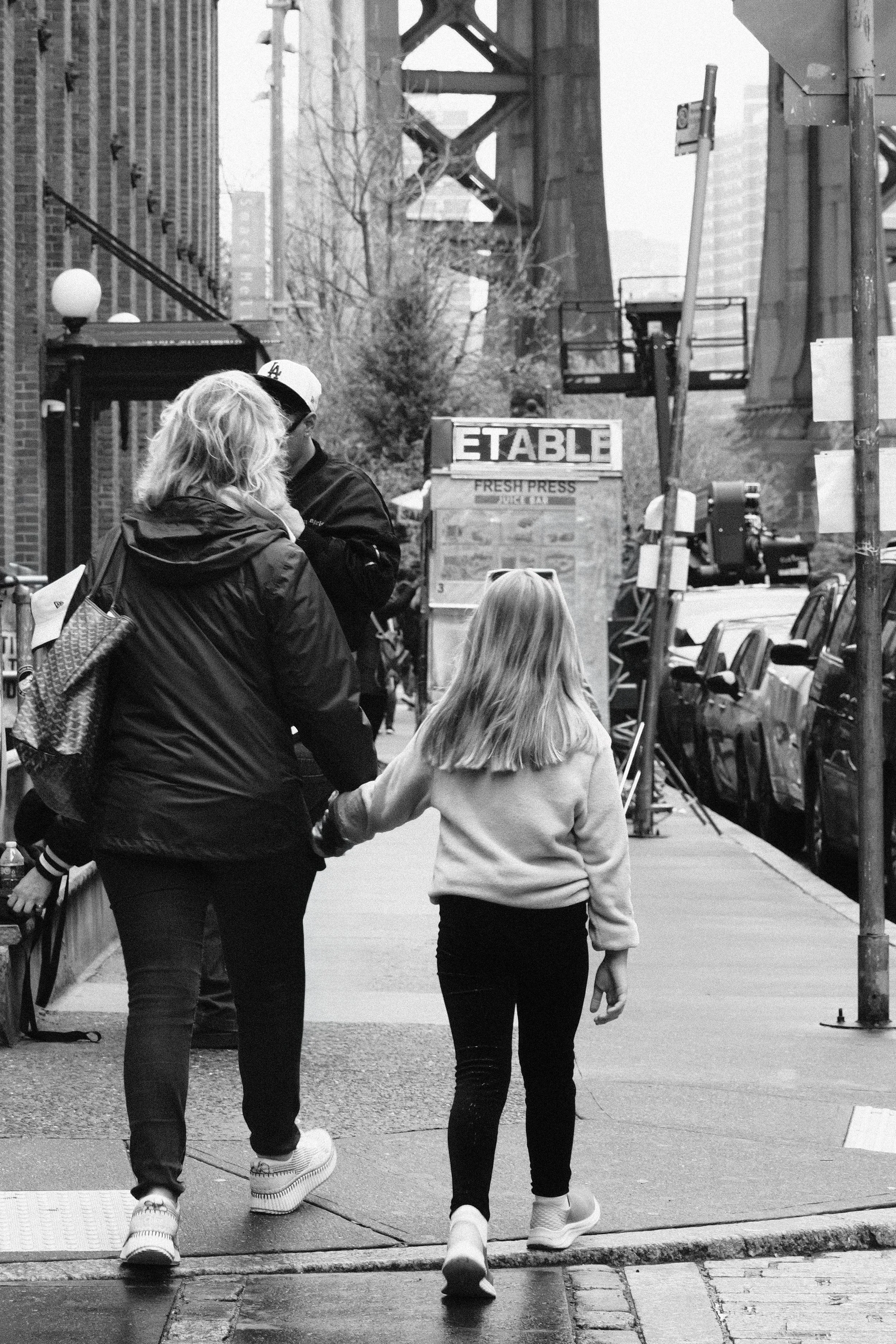 A woman and a young girl hold hands while walking down a city sidewalk, with urban buildings, parked cars, and bridge in the background in black and white.