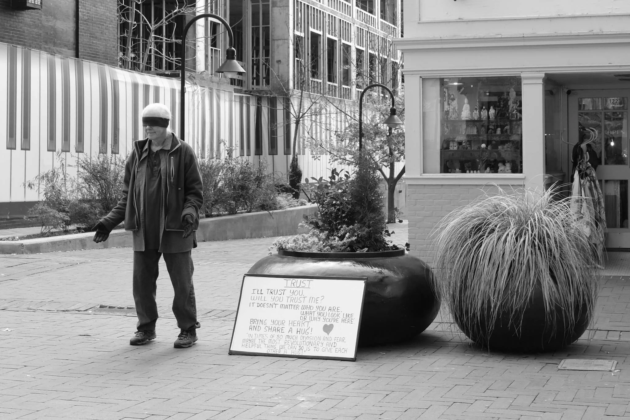 A grayscale photo of a person walking on a city street, wearing a blindfold and gloves, with a large plant and a small patch of shrubs nearby. A handwritten sign leaning against the plant reads about trust and sharing a hug.