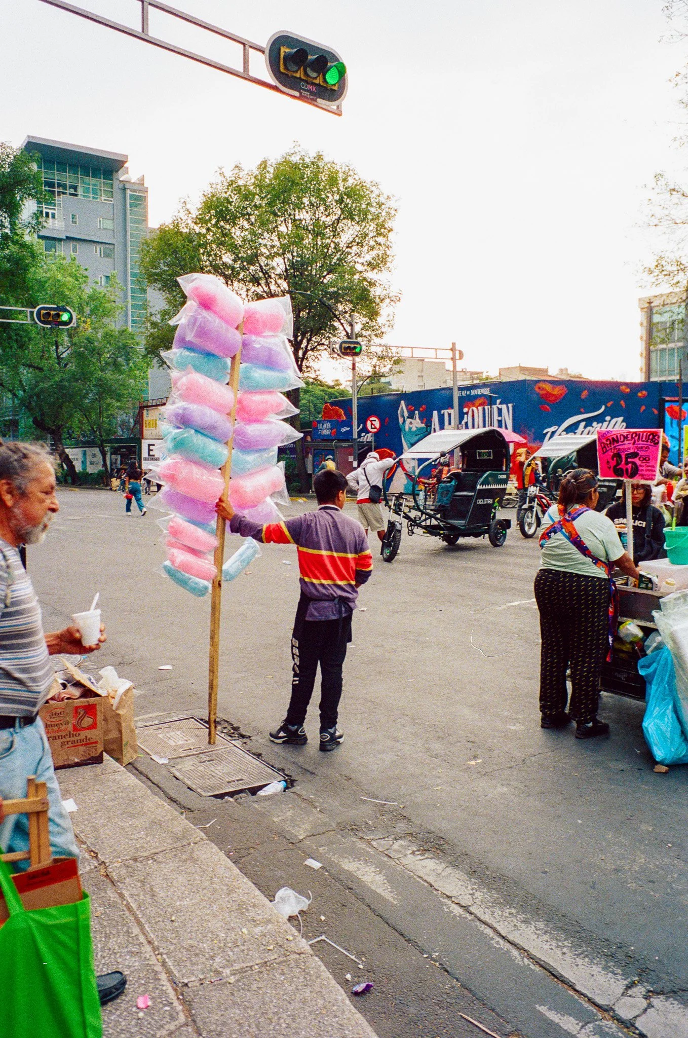 Street scene in Mexico City with cotton candy vendor, pedestrians, and rickshaws, with traffic lights and buildings in the background.