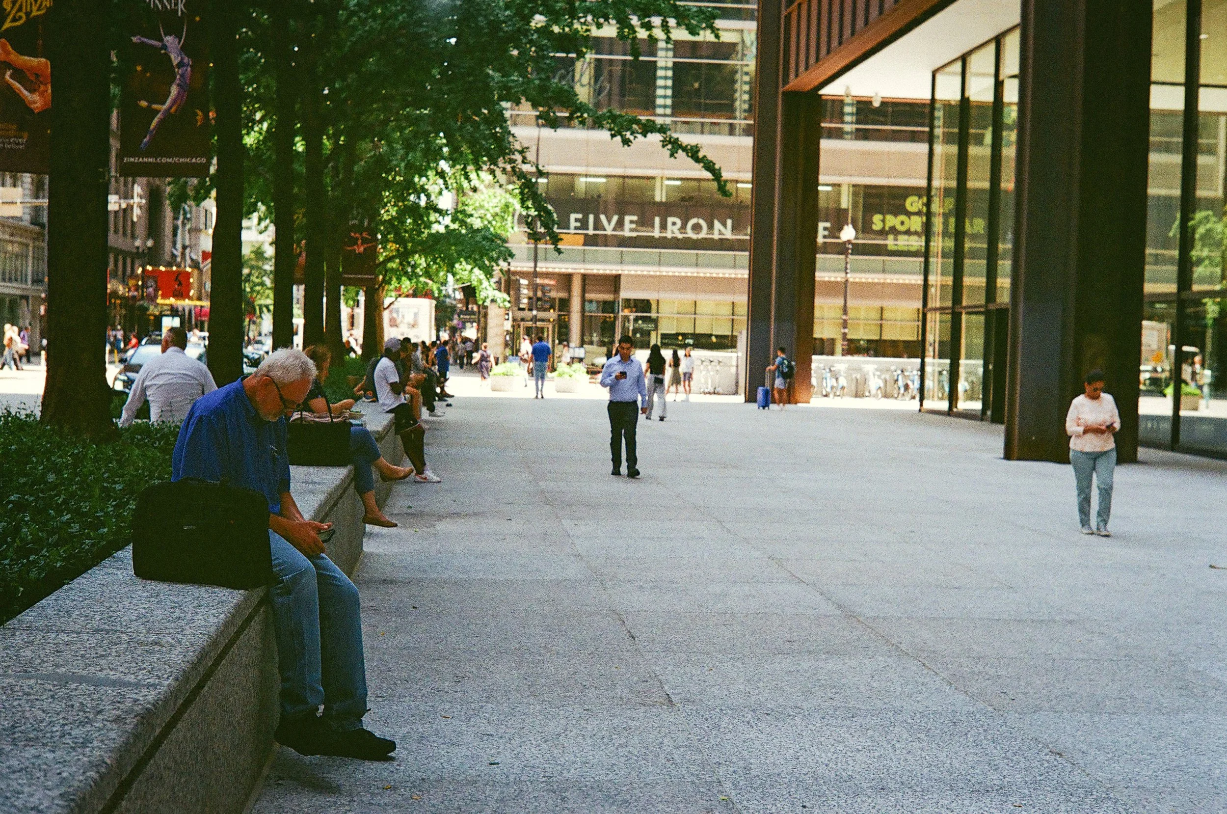 City sidewalk scene with people sitting and standing, modern glass building in the background, trees lining the sidewalk, and a sign reading 'FIVE IRON' on the building.