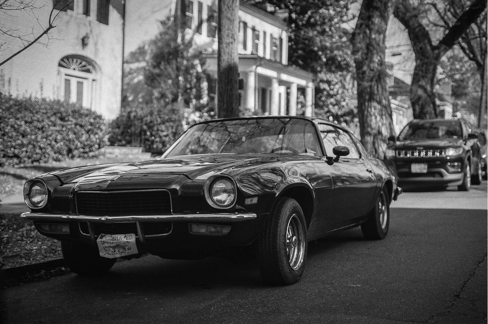 A vintage black sports car parked on a residential street in black and white, with modern cars and houses with porches and trees in the background.