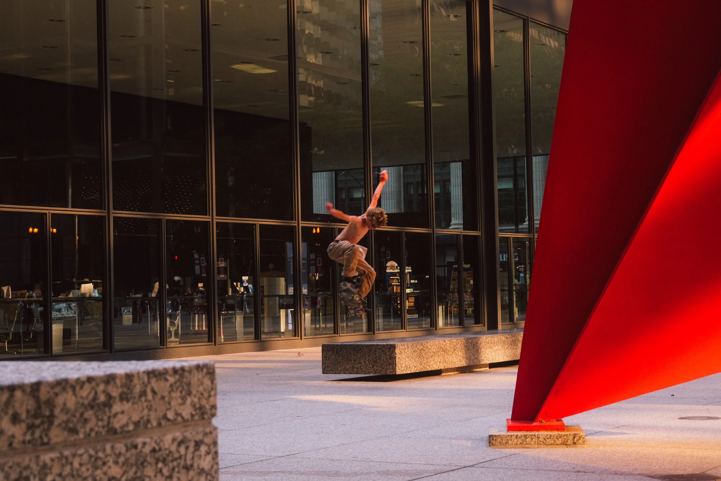 A young man skateboarding in front of a modern glass building with red architectural structure on the right side, during sunset.