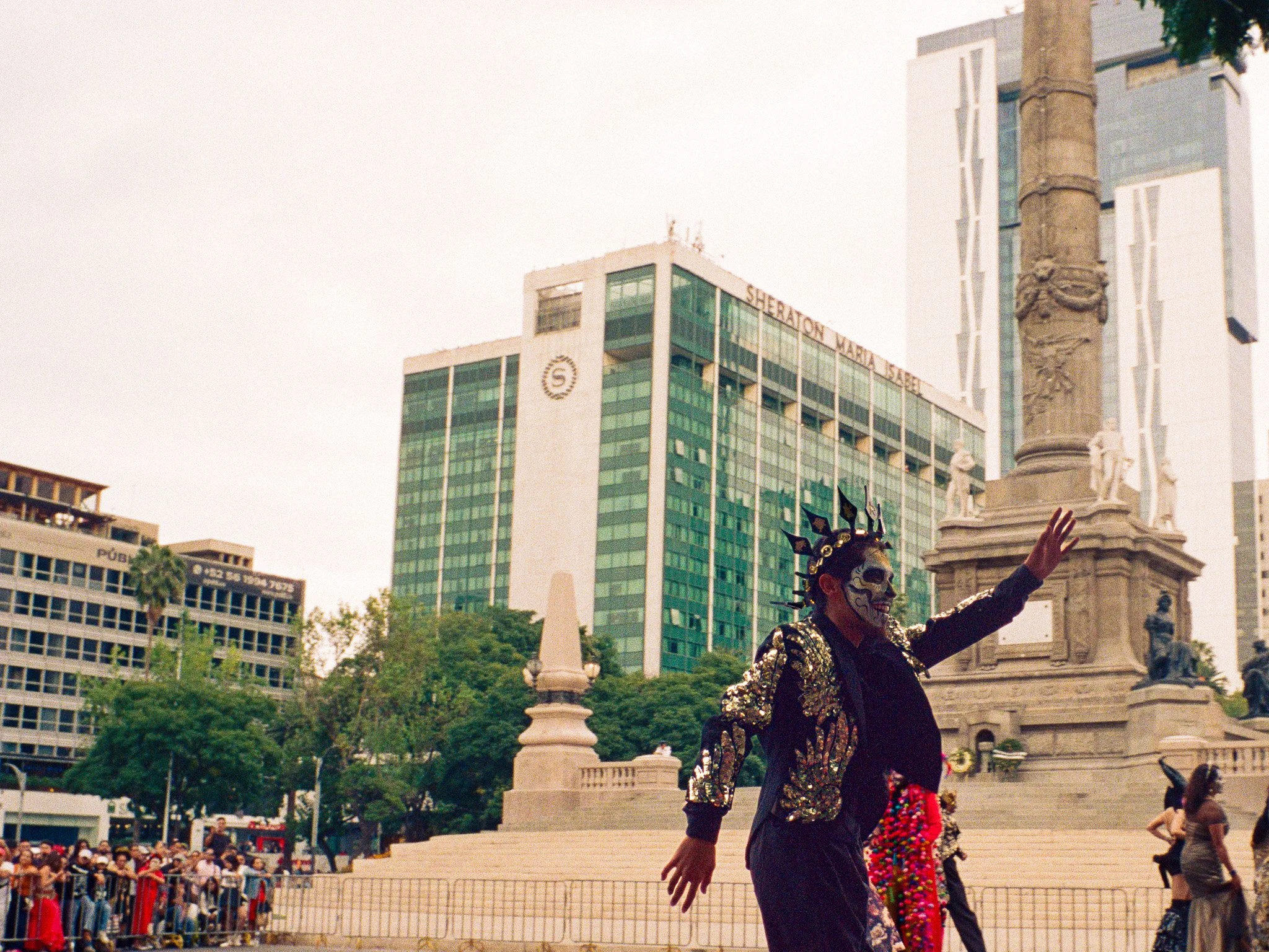 Person dressed as a Día de los Muertos skeleton, wearing a decorative skull mask and black jacket with gold embroidery, celebrating in a public square with a monument and tall buildings in the background.