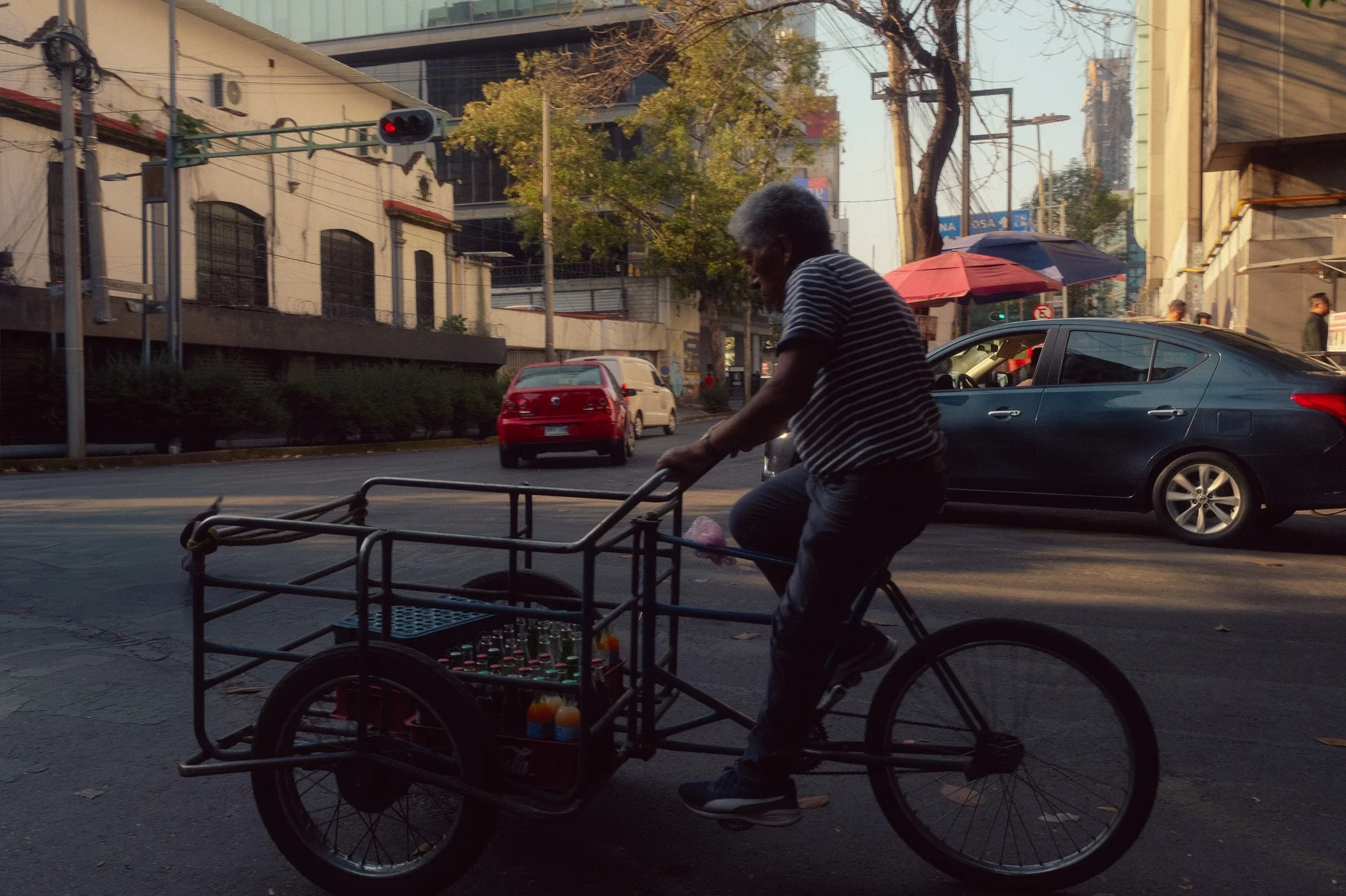 An elderly man riding a bicycle with a cart attached to carry bottles on a city street, during late afternoon or early evening.