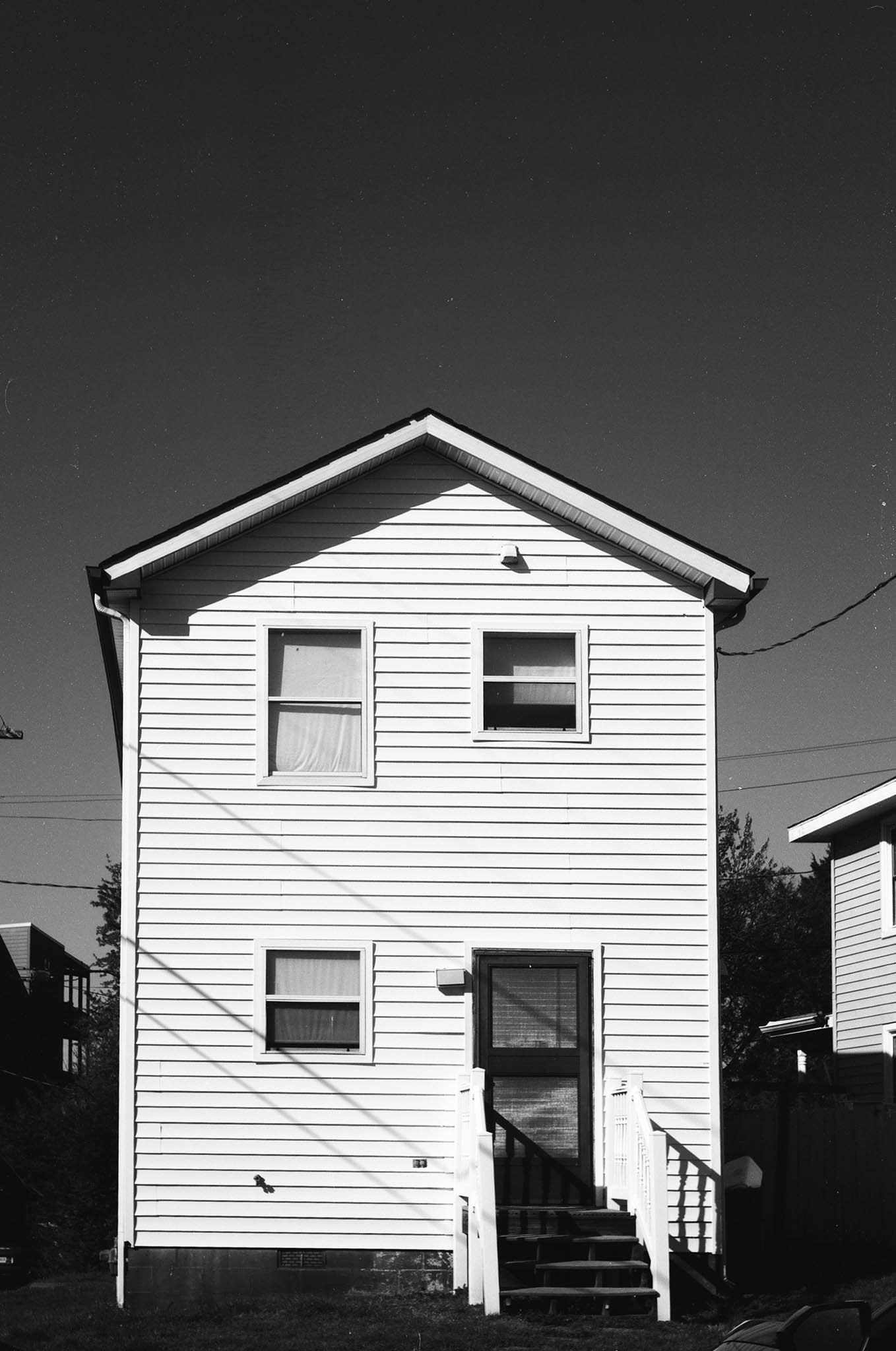 A two-story house with white siding, two windows on the upper floor and two on the lower, with stairs leading to a door. The house is in black and white with a clear sky above.